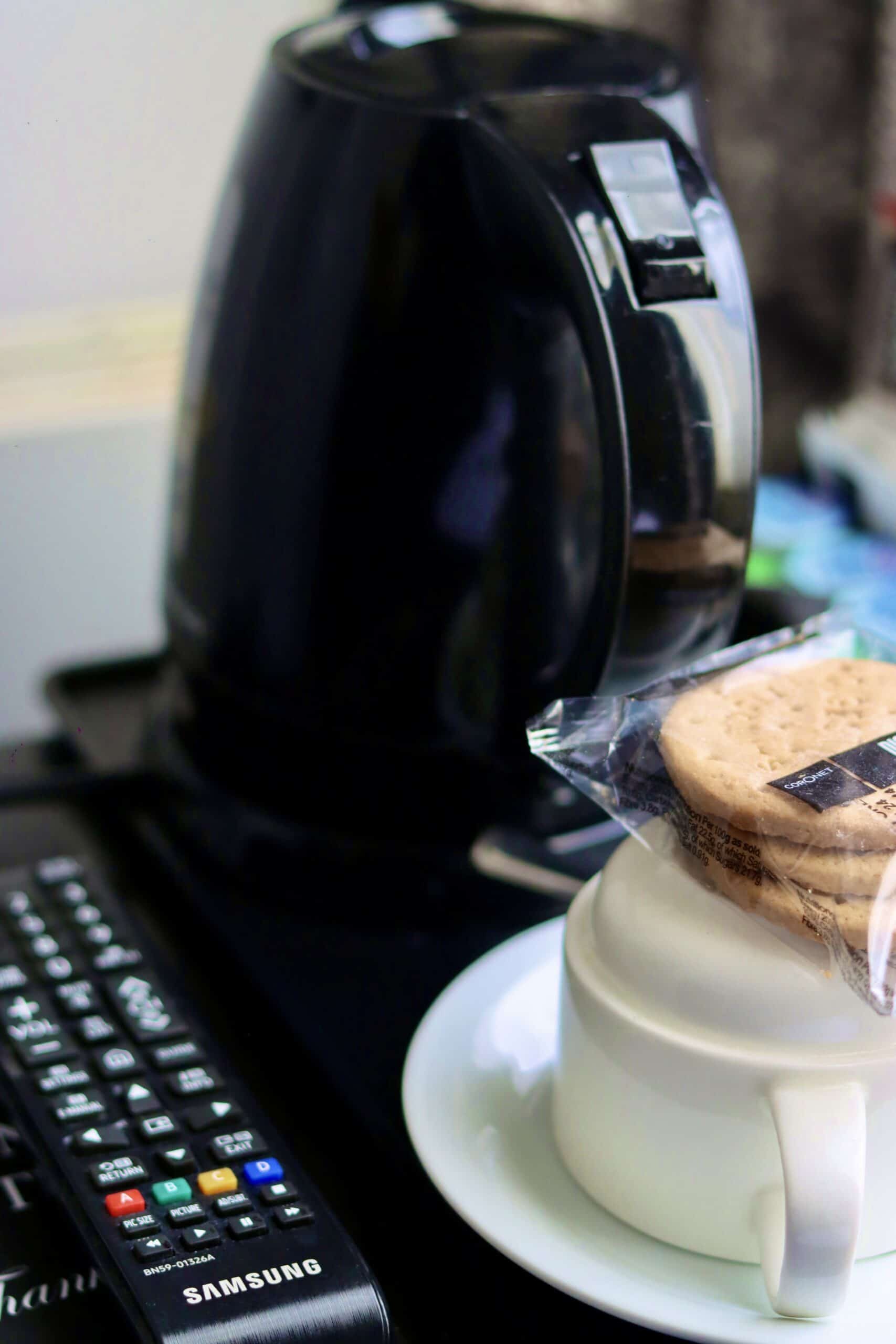 A black electric kettle sits on a tray next to a remote control. In the foreground, there is a white cup with a saucer and a pack of biscuits placed on top of the cup.