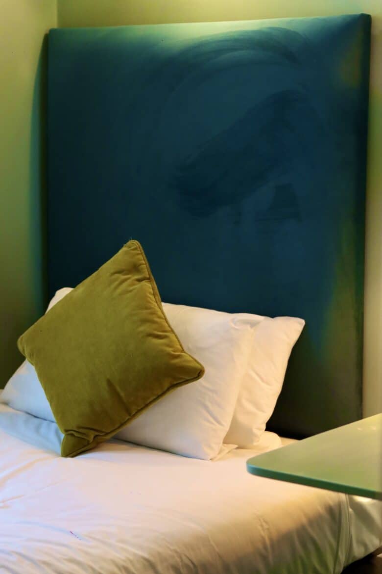 A neatly made bed with white linens is seen with two white pillows and one green pillow propped up against a large padded teal headboard. The room has a minimalist design with a green wall and a small glass table edge visible in the foreground.