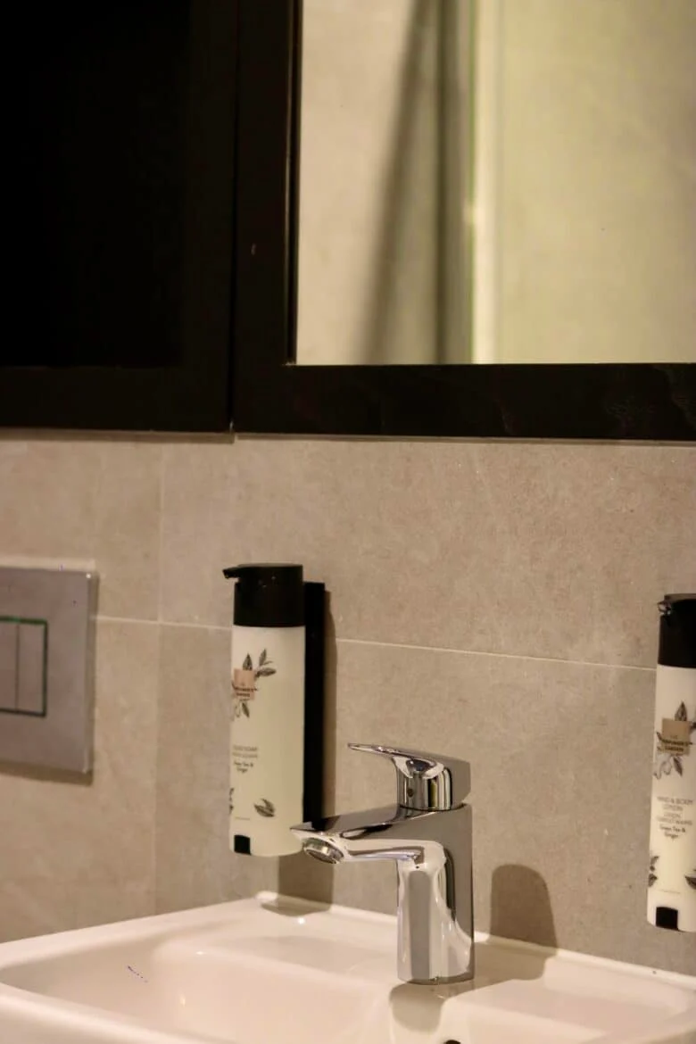 A modern bathroom sink with a chrome faucet is set against a tiled wall. Above the sink, there is a large mirror with a black frame. Wall-mounted soap dispensers are placed on either side of the sink.