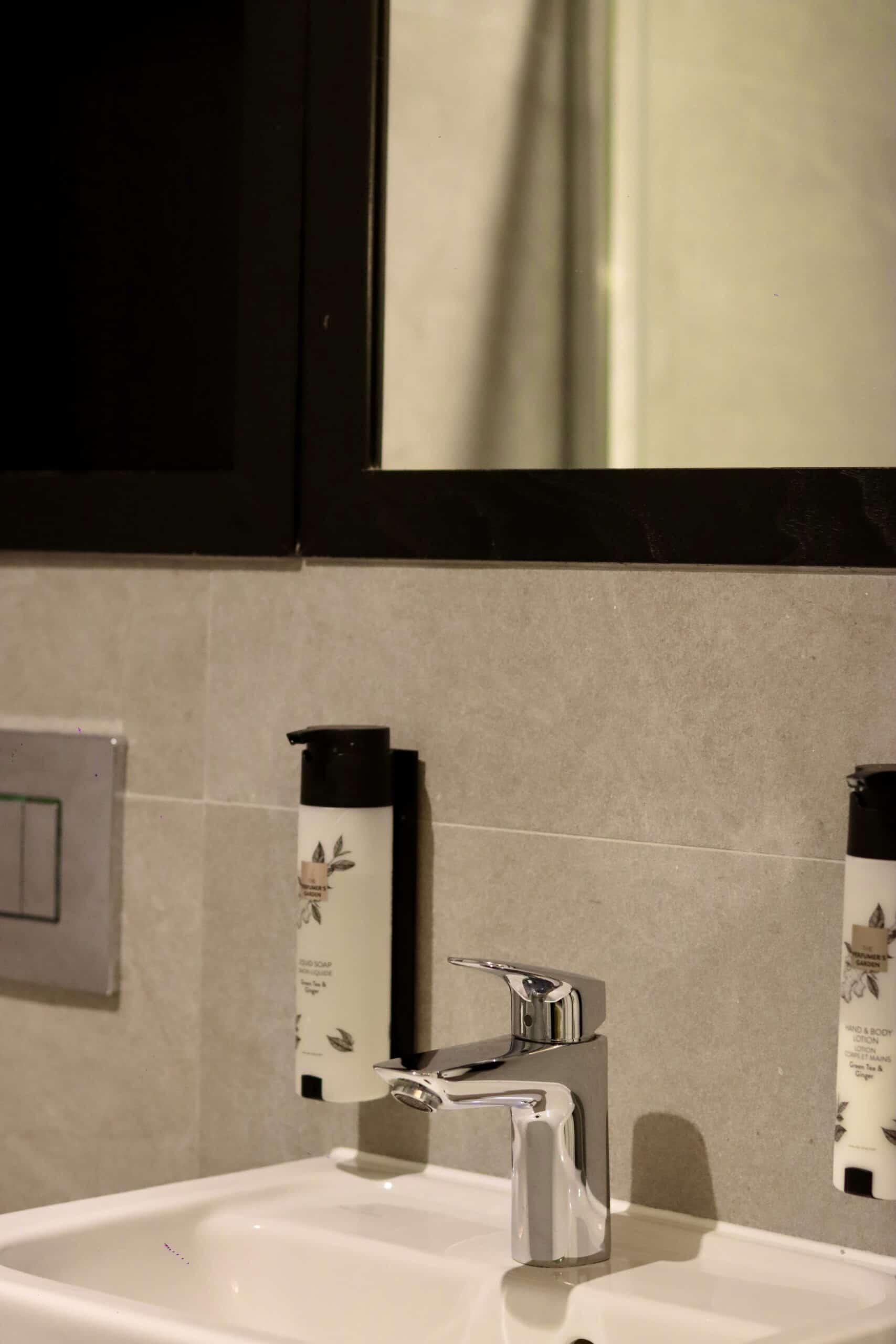 A modern bathroom sink with a sleek, chrome faucet. Above the sink is a large mirror with a black frame. On either side of the sink are two bottles of lotion or soap mounted on the wall. The walls are tiled in a light gray color.