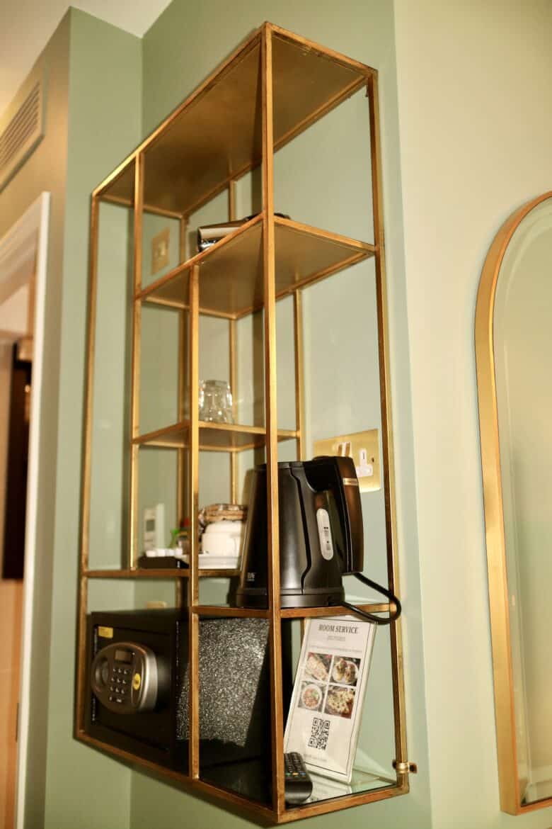 A wall-mounted shelf made of gold-colored metal holds a variety of items, including a safe, a kettle, a glass, and a framed room service menu. The shelf is positioned near a light green wall and a doorway.