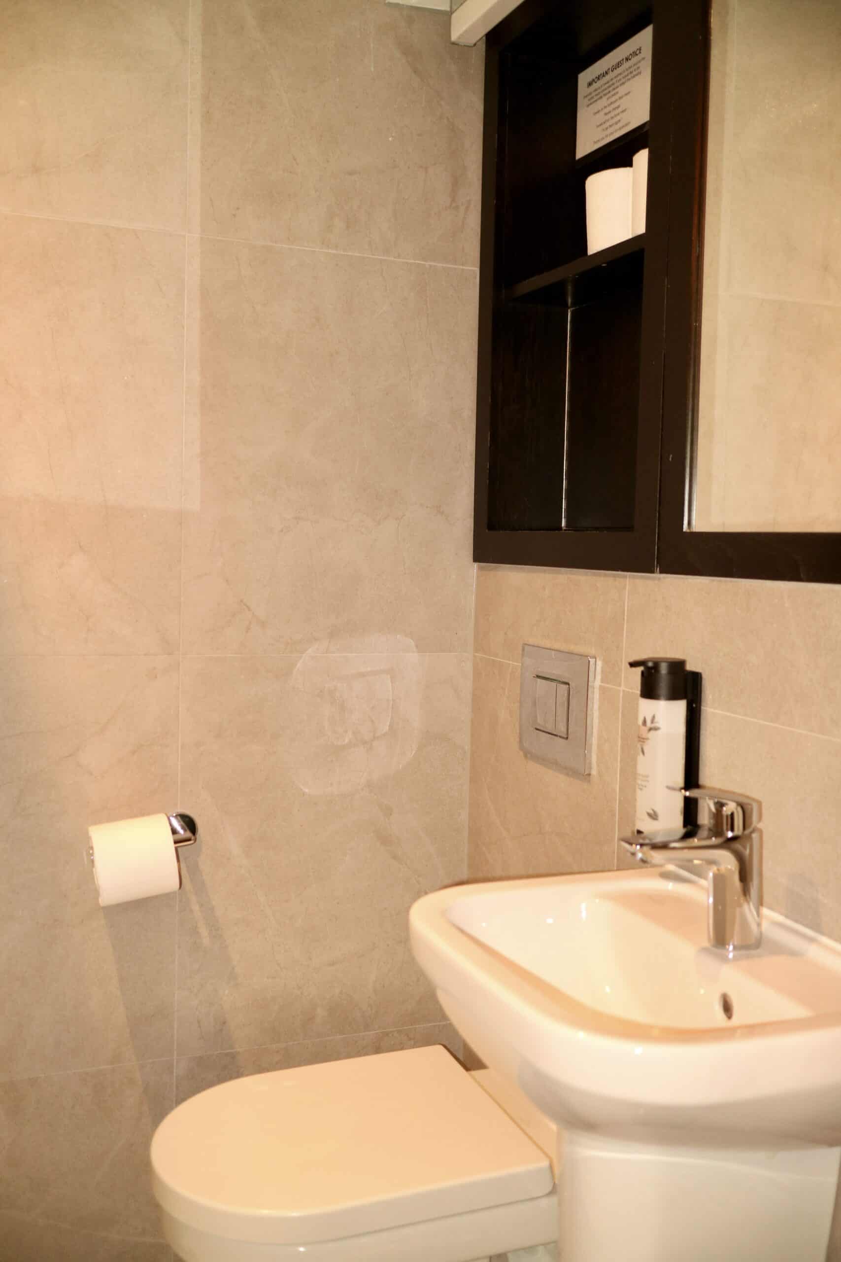 A modern bathroom featuring a wall-mounted toilet with a roll of toilet paper on a nearby holder, a white sink with a chrome faucet, and a dark brown shelving unit above the sink containing a small sign and decor items. The walls are covered with light beige tiles.