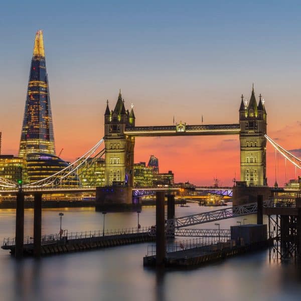 Tower Bridge in London illuminated at dusk with the Shard skyscraper in the background. The sky shows hues of orange, pink, and blue, reflecting off the River Thames, creating a picturesque and serene evening scene — perfect for visitors staying in nearby hotels in London.