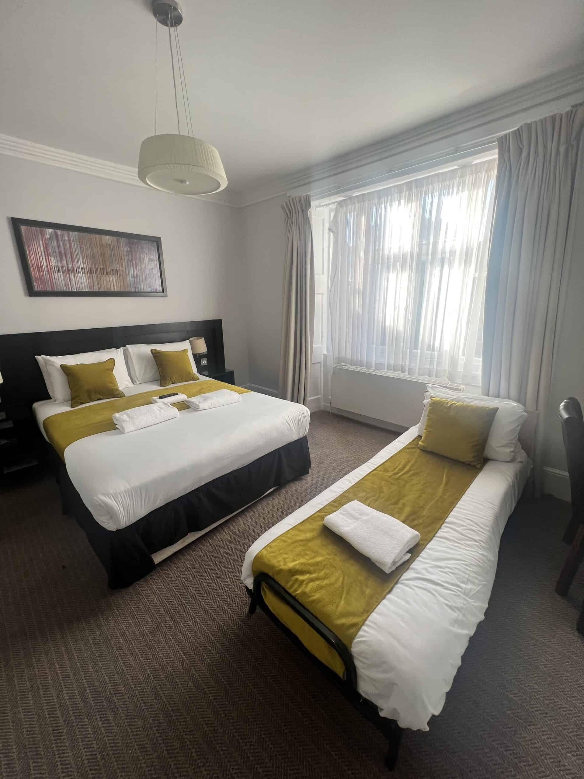 A bright hotel room with a large window features a double bed and a single bed, both adorned with white linens and yellow accents. The room includes a modern light fixture, wall art above the double bed, and neatly folded towels on each bed.