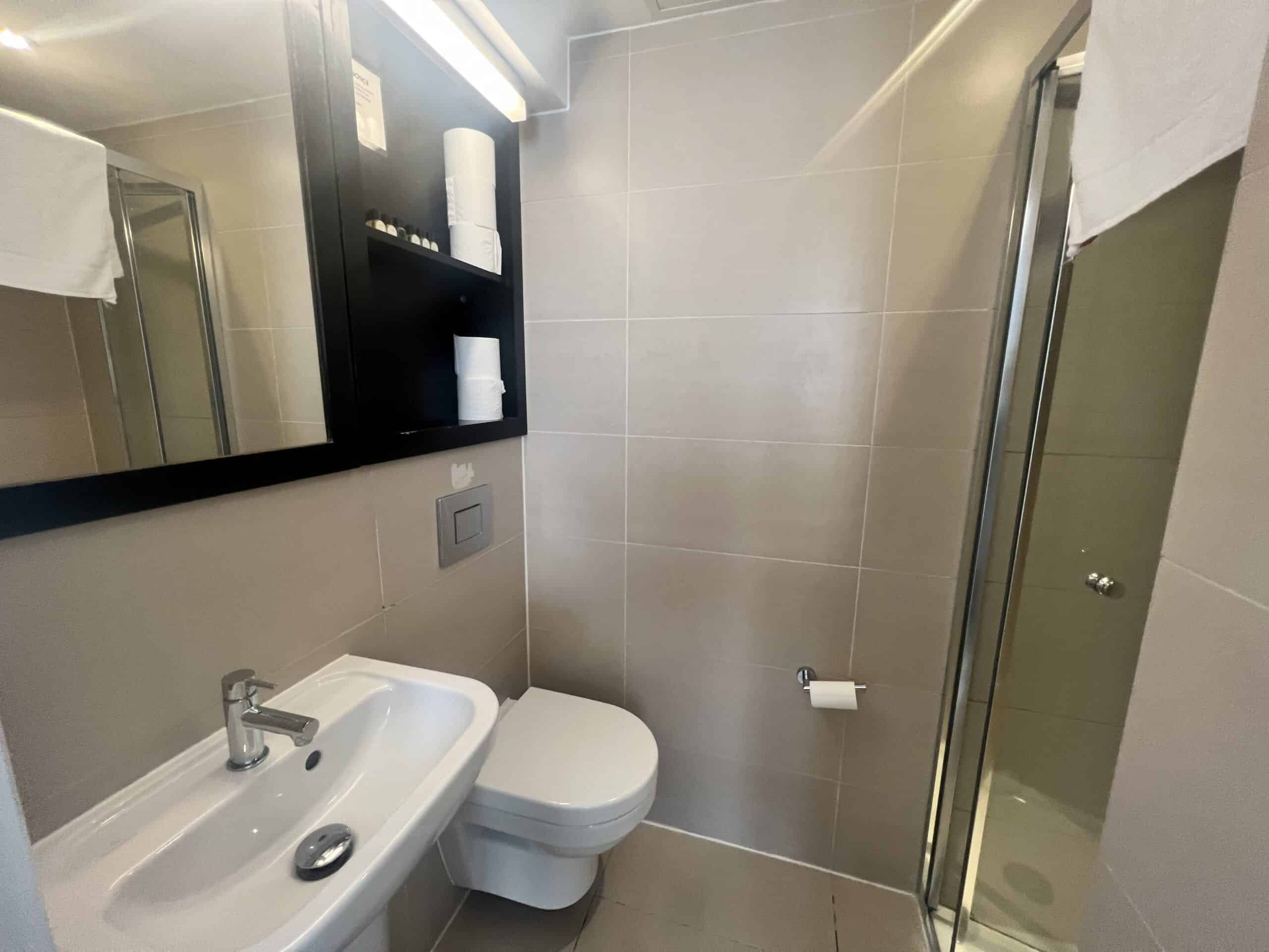 A modern bathroom with beige tiles, a white sink with a silver faucet, a toilet, a wall-mounted black cabinet with toiletries, a mirror, and a shower with a glass door. A toilet paper roll is mounted on the wall next to the toilet.