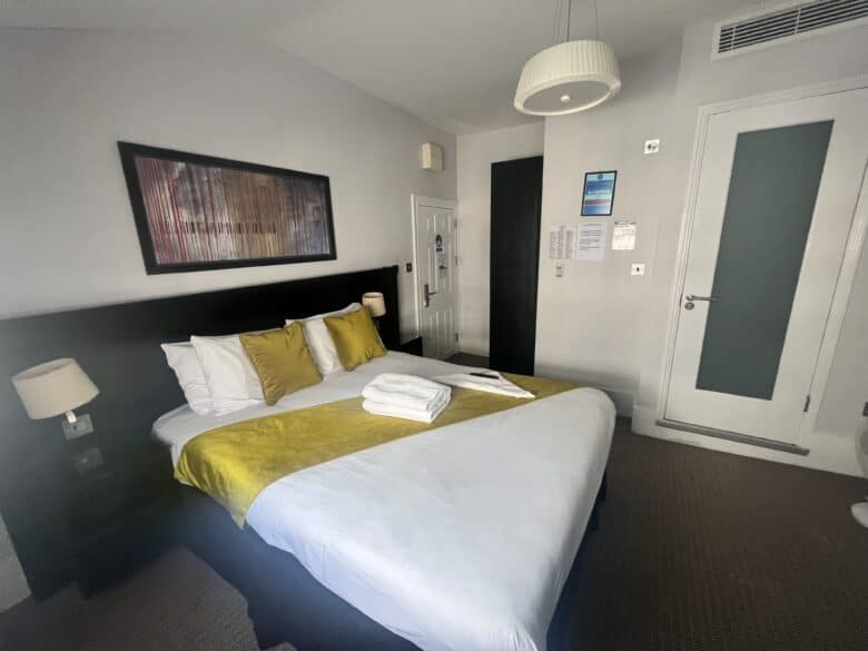 A modern hotel room with a neatly made bed featuring yellow and white bedding. Two bedside tables with lamps are on either side. A large abstract painting hangs above the bed. The room has a white door and a frosted glass door. Towels are folded on the bed.