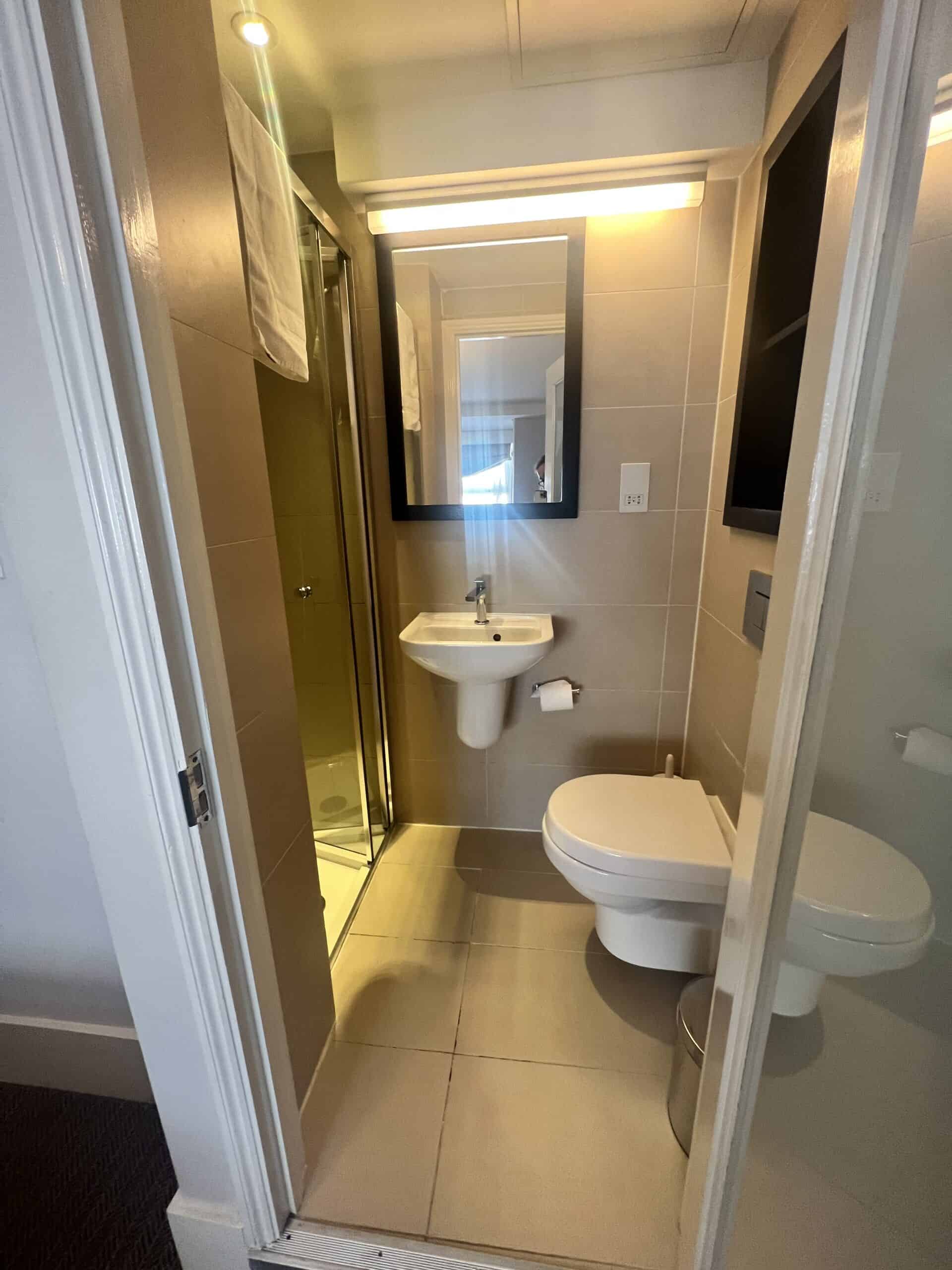 A compact bathroom featuring a wall-mounted sink, a mirror with overhead lighting, a floating toilet, a glass-enclosed shower, and beige tiled walls and floor. The doorway is partially visible, with a towel hanging on the shower door.