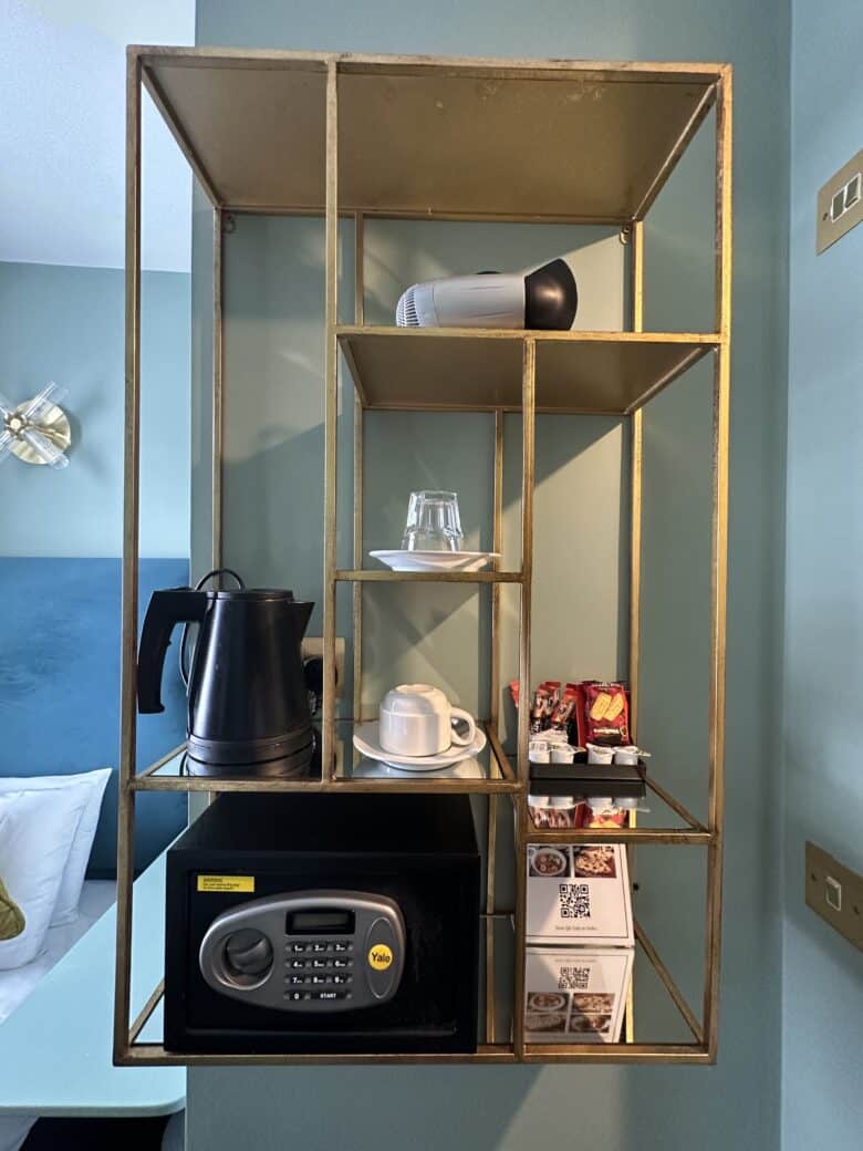 A gold-colored wall shelf holds a black electric kettle, a flashlight, an upside-down glass, a white cup with saucer, a small black safe, packets of instant coffee, and multiple restaurant brochures. A bed with a blue headboard is partially visible on the left.