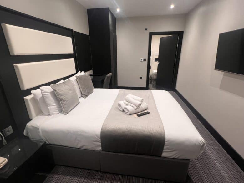 A modern hotel room with a neatly made king-sized bed featuring white and gray linens and several pillows. Two white towels are folded on the bed along with a TV remote. The room has dark furniture, including a desk and nightstand, with a doorway leading to another area.