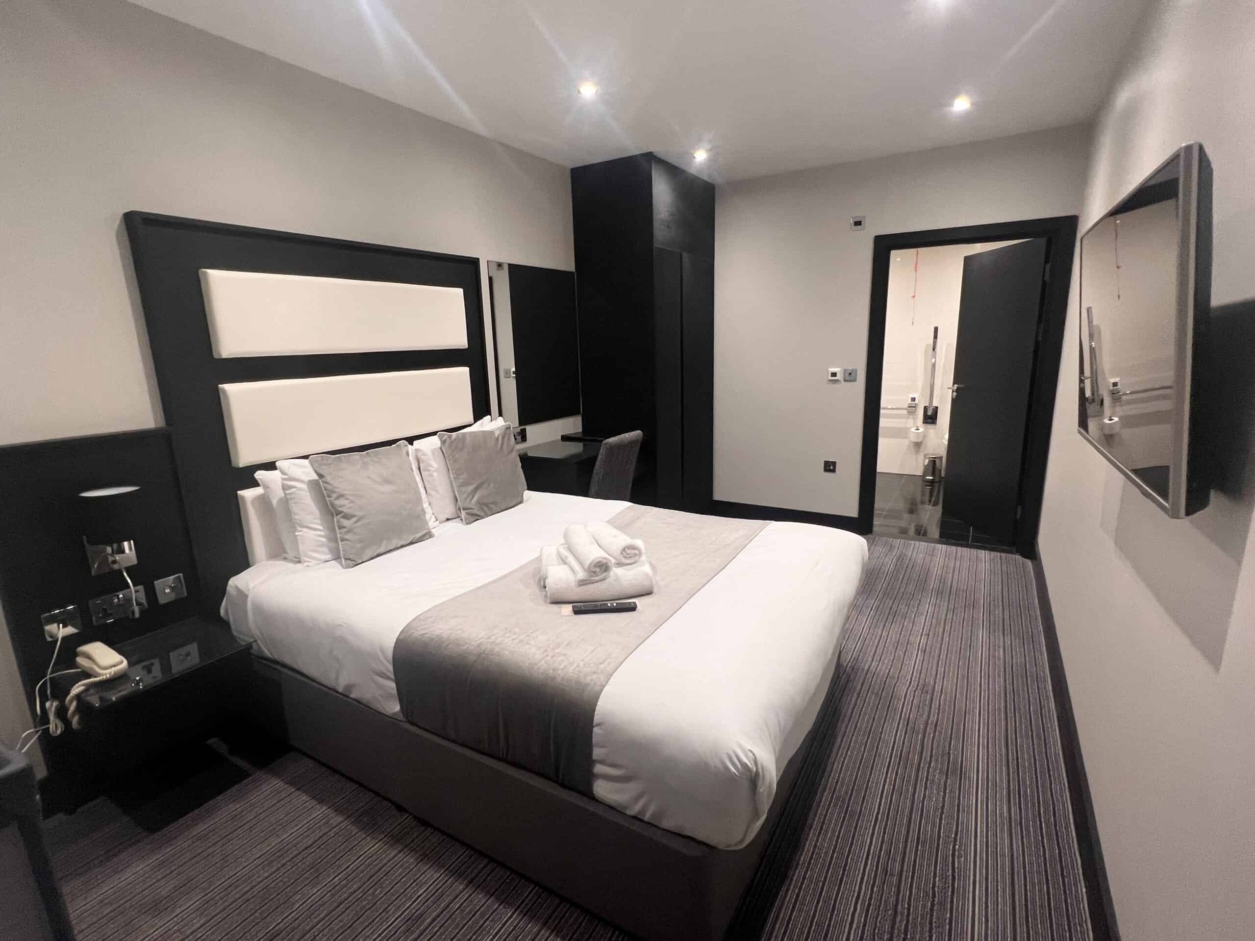 A modern hotel room features a neatly made double bed with gray and white bedding and two pillows, topped with folded towels. There is a wall-mounted TV to the right, a desk with a chair, and an open door leading to a bathroom at the end of the room.