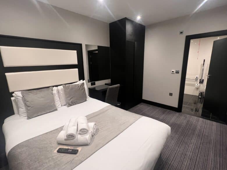 A neatly arranged hotel room with a double bed, gray and white bedding, and four pillows. Towels and a TV remote rest on the bed. A desk with a chair and a large mirror are visible. The door to the bathroom is open, revealing a glimpse of the facilities inside.