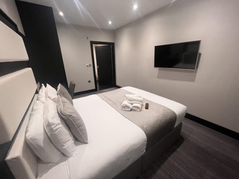 A modern hotel room with a double bed, white and gray linens, and multiple pillows. A neatly folded towel and a remote control are placed on the bed. The room features a wall-mounted flat-screen TV, neutral-colored walls, and carpeted flooring.