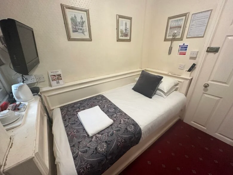 A small, cozy hotel room features a neatly made single bed with a white towel on top, and adorned with a dark patterned runner and cushions. The room includes a wall-mounted TV, framed pictures, and basic amenities like a kettle and tea supplies on a side shelf.