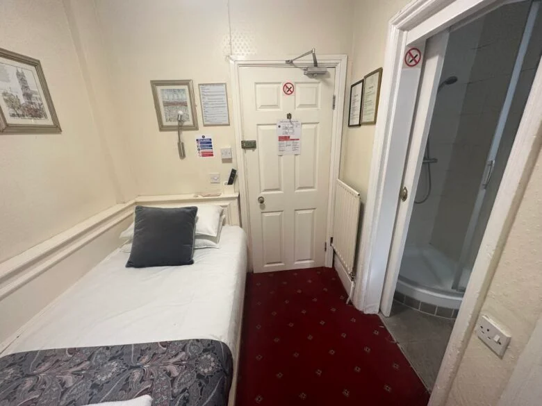 A small hotel room with a single bed, white linens, and two gray pillows. The room has a red carpet and cream-colored walls adorned with framed pictures. An en-suite bathroom with a glass shower door is visible. Various signs and notices are posted on the main door.