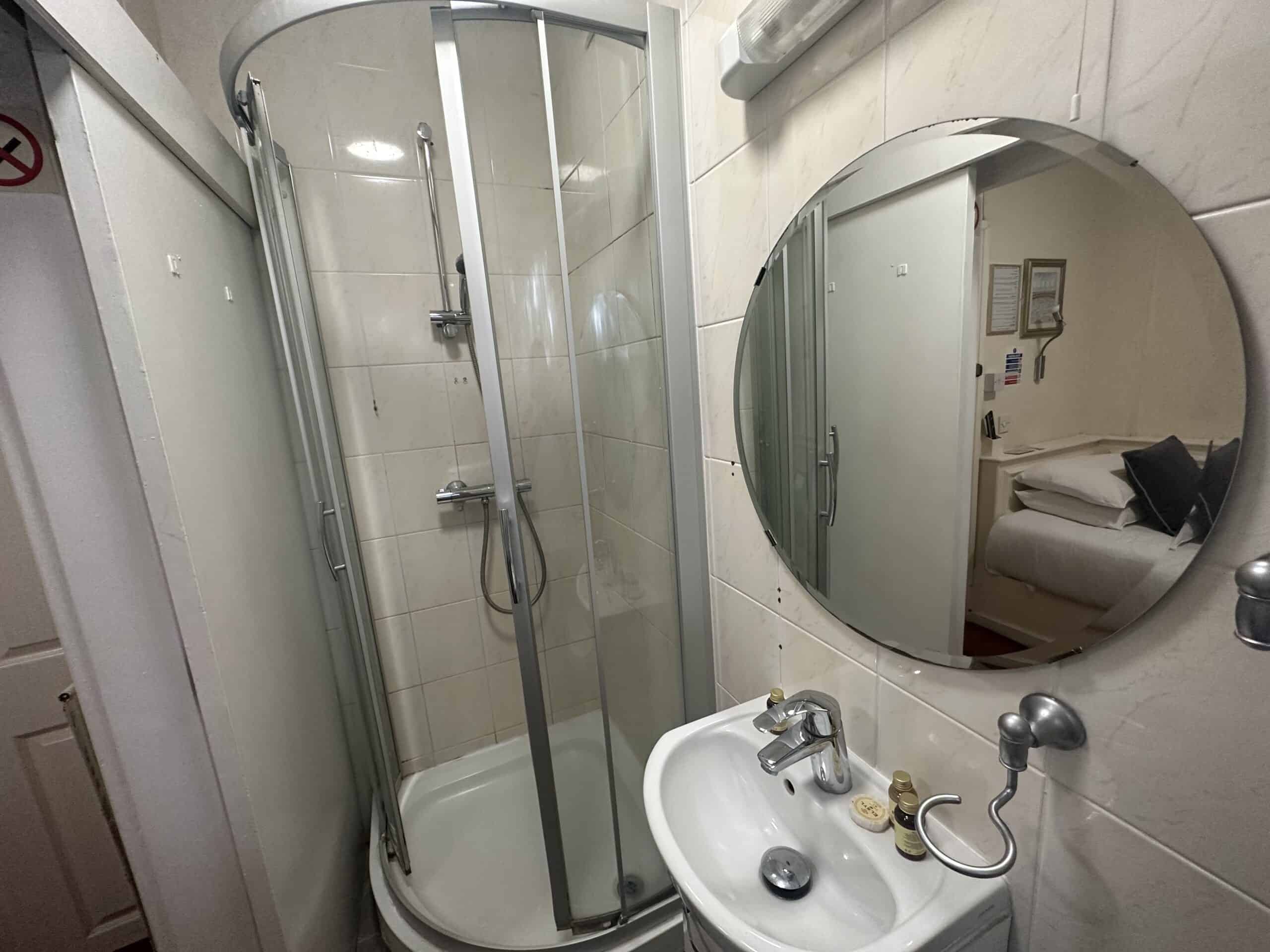 A small bathroom featuring a curved glass shower with metal frame, a round mirror above a white sink, and a wall-mounted light. The shower has a handheld showerhead, and the sink has small toiletries. A glimpse of a bedroom can be seen in the mirror's reflection.