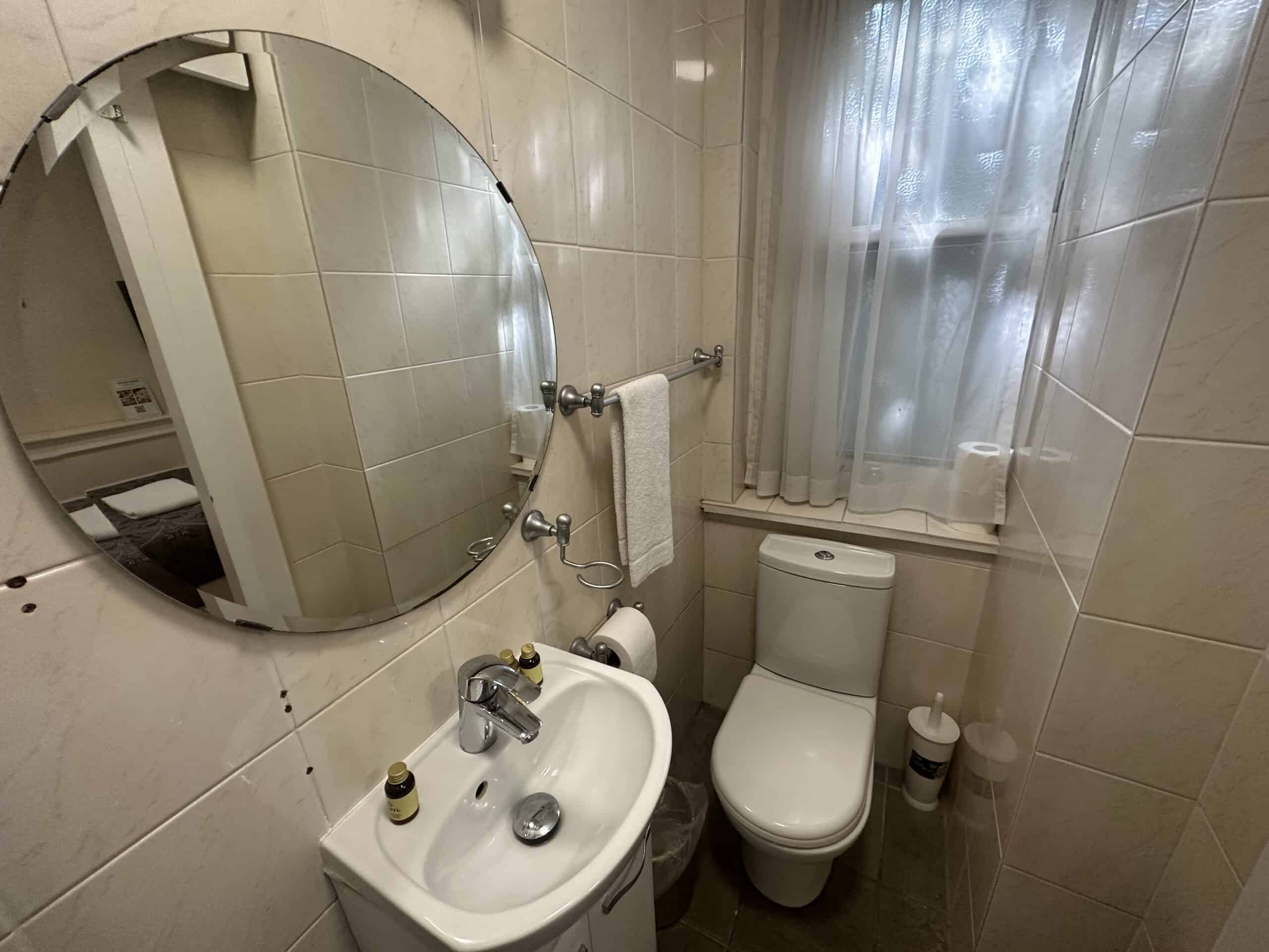 A small bathroom with beige tiled walls features a round mirror above a compact white sink with toiletries, a towel holder with a white towel, and a modern white toilet. A frosted window allows natural light to filter in, and a toilet paper holder is mounted nearby.