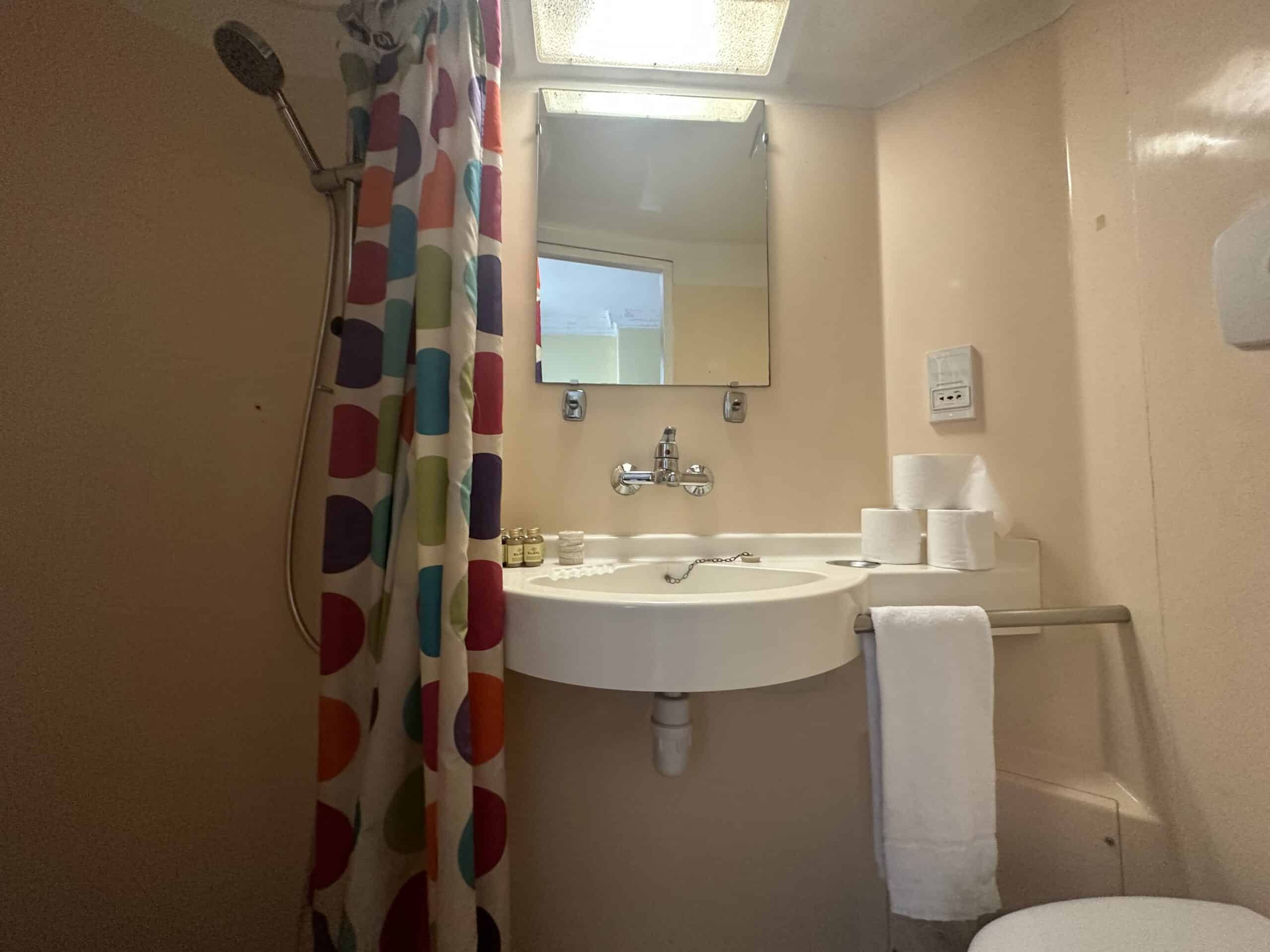 A small bathroom features a shower on the left with a colorful curtain, a circular white sink with toiletries and a towel, a mirror above the sink, and a toilet next to it on the right. The walls are light-colored, and a ceiling light illuminates the space.