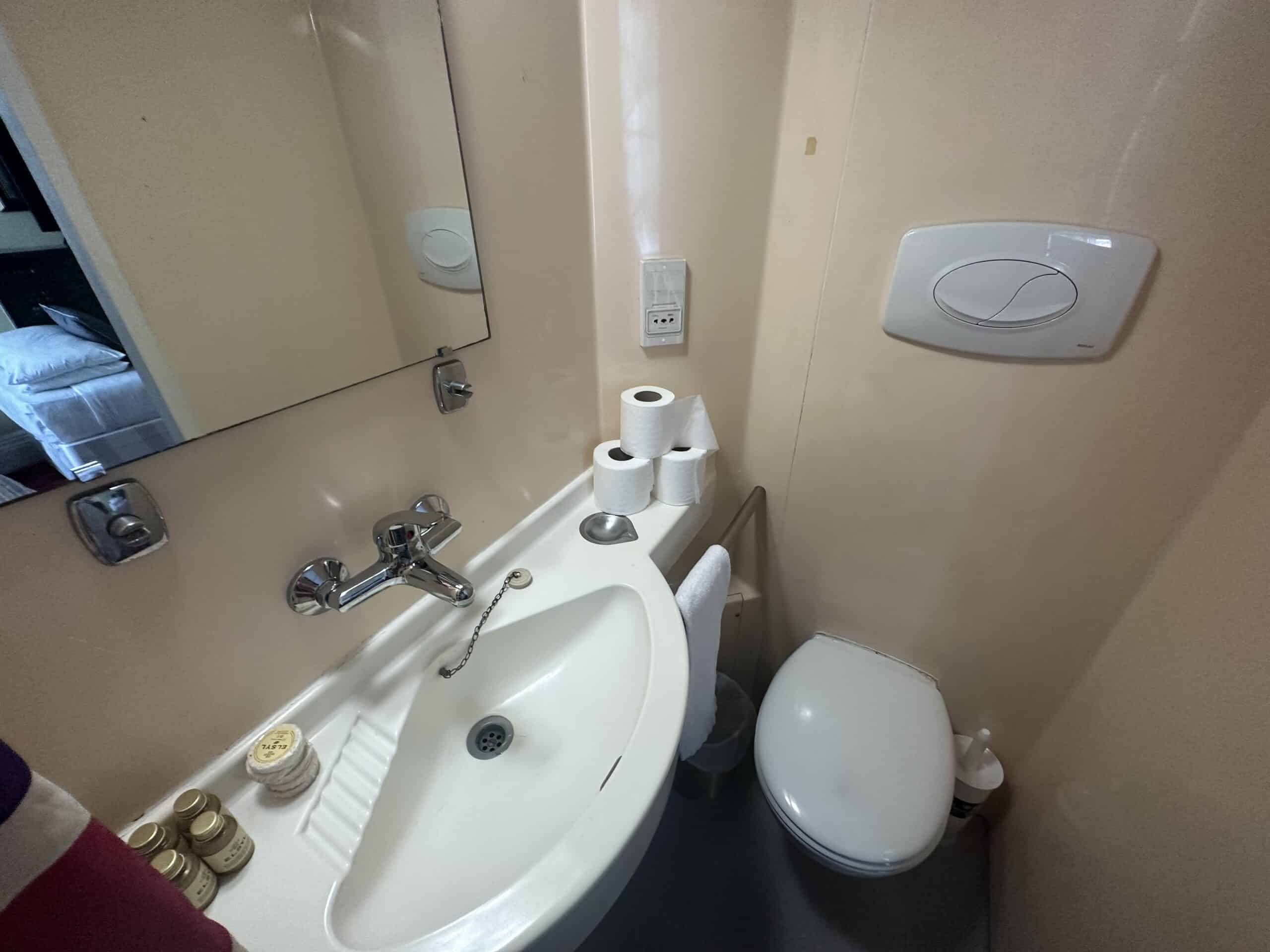 A compact bathroom with a sink, mirror, toilet, and toiletries. The sink has a single faucet and holds several bottles and soaps. Above the toilet, mounted on the wall, is a white flush button. Three rolls of toilet paper are stacked beside the sink.