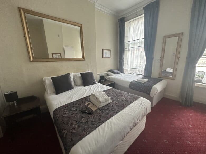 A hotel room features two neatly made beds with white linens and decorative dark runners. One bed has a mirror above it. Dark curtains frame a large window, and a full-length mirror leans against the wall. There is a nightstand with a lamp between the beds.