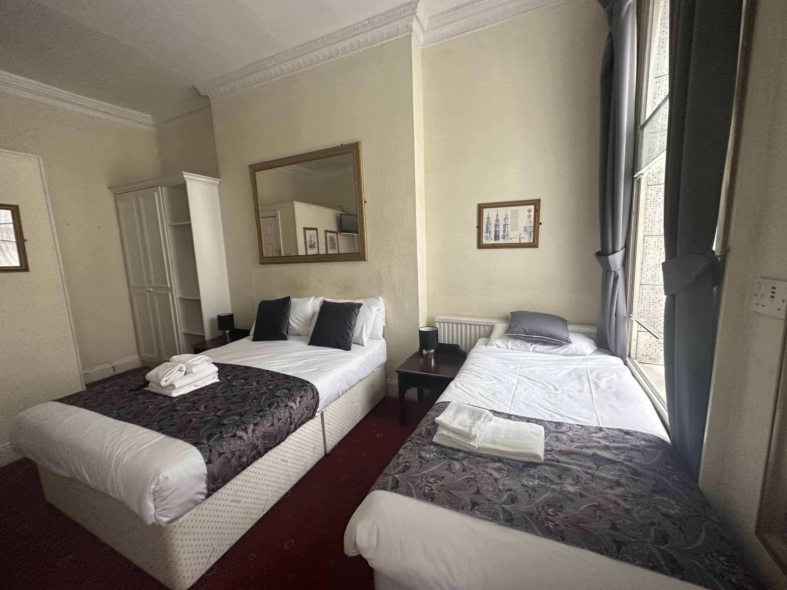 A hotel room featuring two neatly made beds with dark patterned runners and white linens. Each bed has a set of folded towels. A framed mirror hangs above one bed. The room also includes a wardrobe, a bedside table, and a large window with curtains partially drawn.