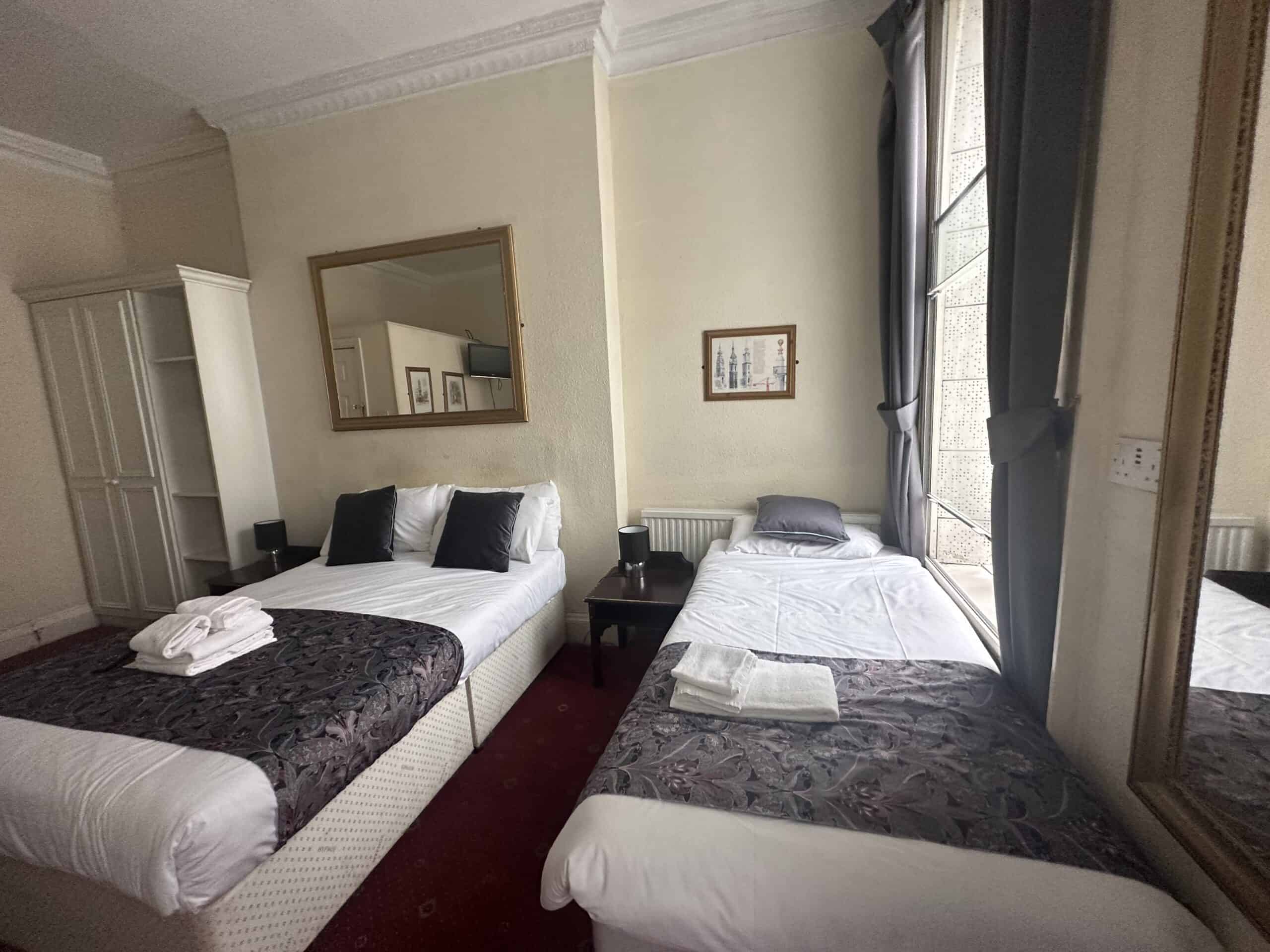 A hotel room featuring two beds: a double bed on the left with white linens and gray decorative pillows, and a single bed on the right with white linens. The room includes a large mirror above the double bed, a wardrobe, and a window with curtains. A folded towel is placed on each bed.