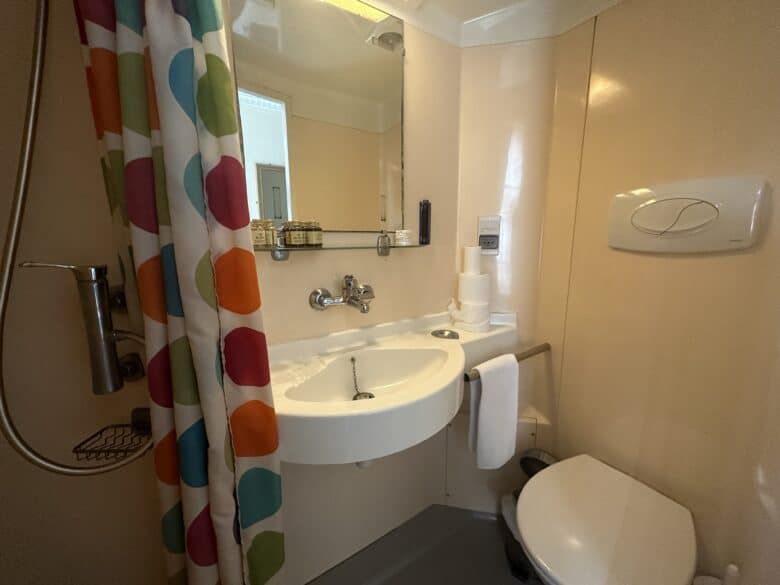 A small bathroom featuring a sink with toiletries on the counter, a round mirror above it, and a white toilet next to the sink. The shower area has a colorful polka dot curtain. The walls are light-colored, creating a clean and compact space.