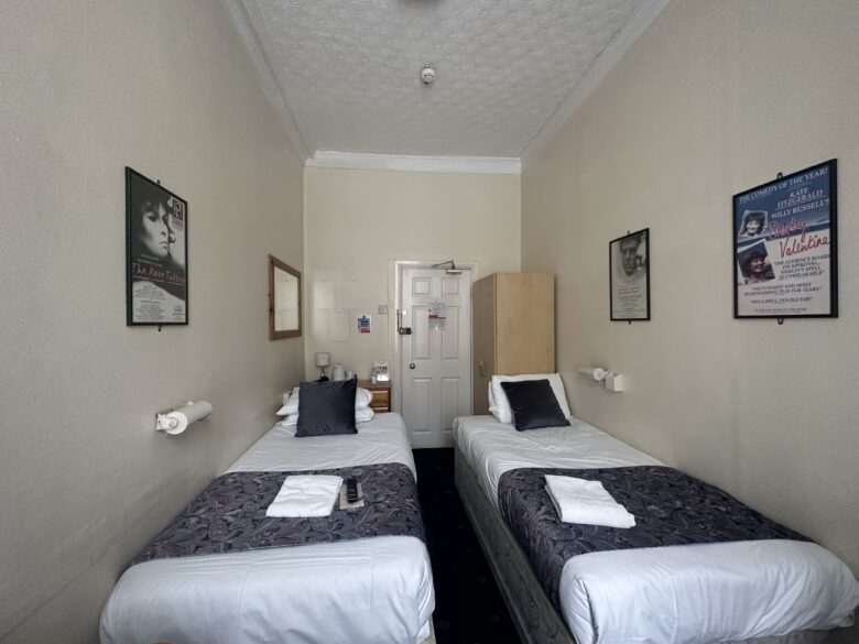 A small hotel room with two single beds side by side. Each bed has white sheets, a dark patterned bed runner, and a folded towel on top. A nightstand with a phone and lamp is between the beds. Posters hang on the walls, and a wooden wardrobe stands near the door.