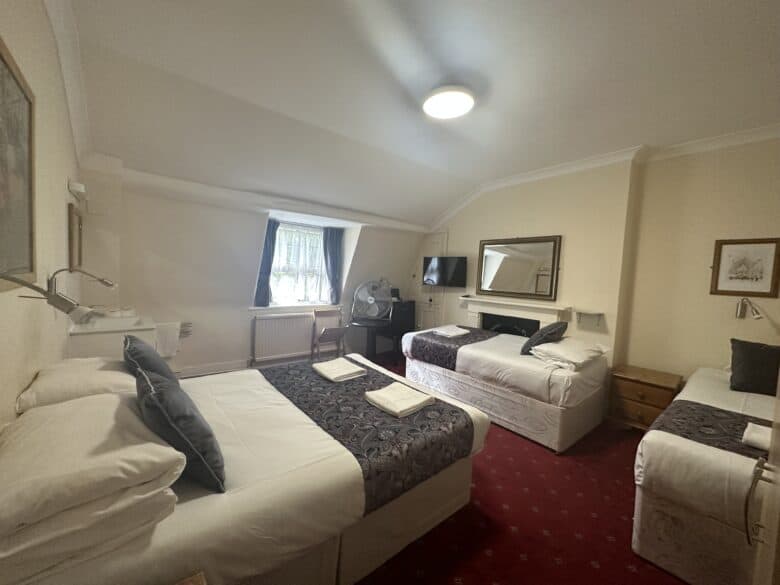 This image shows a cozy hotel room with three beds, each adorned with white linens and dark-colored accent blankets. The room features a red-carpeted floor, a wall-mounted flat-screen TV, and a small fan on a bedside table near a window with curtains.