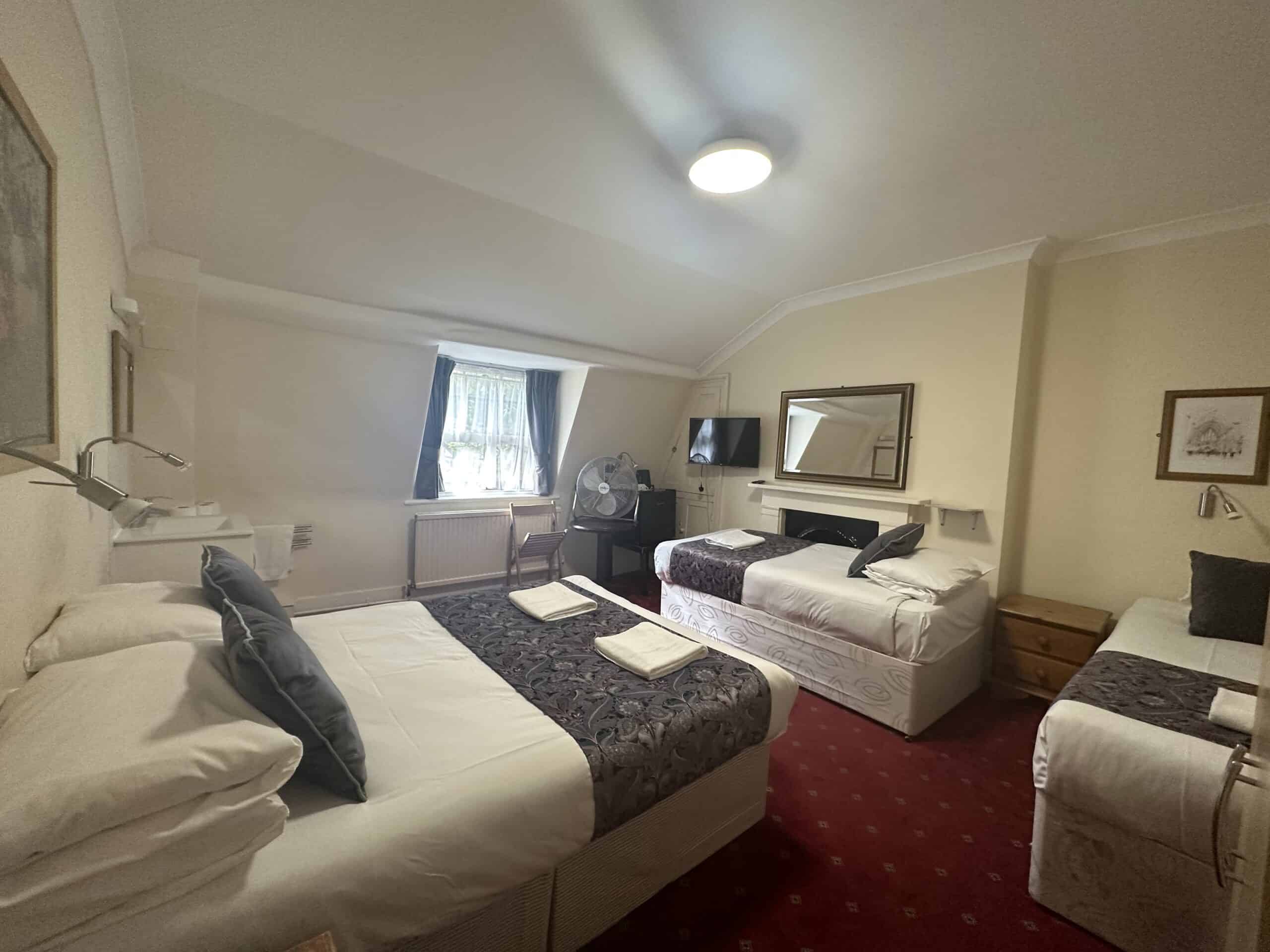 This image shows a cozy hotel room with three beds, each adorned with white linens and dark-colored accent blankets. The room features a red-carpeted floor, a wall-mounted flat-screen TV, and a small fan on a bedside table near a window with curtains.