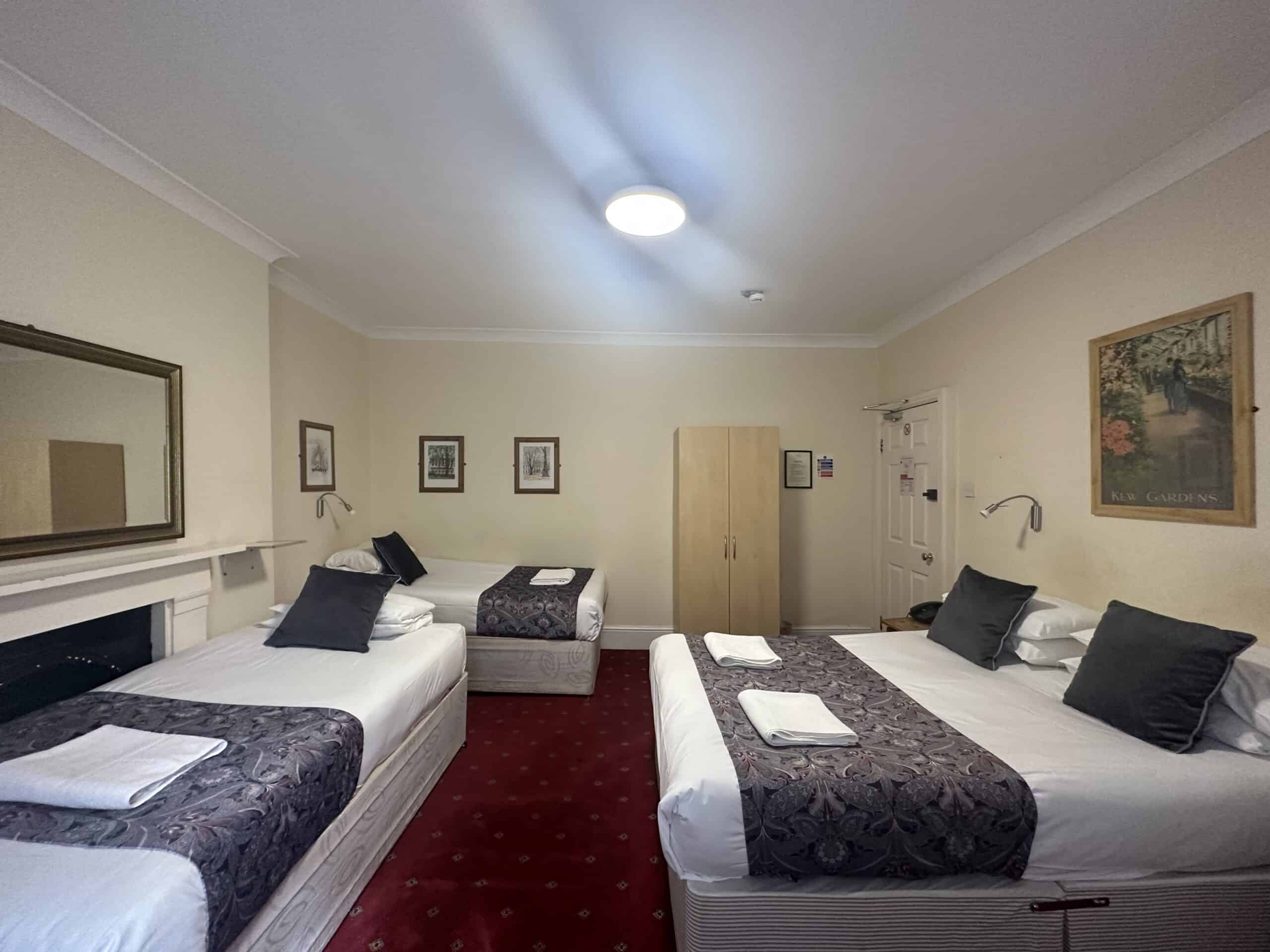 A hotel room with three beds: two singles and one double bed, all with white sheets and dark patterned throws. The room has beige walls, a red carpet, a large mirror above a fireplace, framed artwork on the walls, a wooden wardrobe, and a ceiling light.