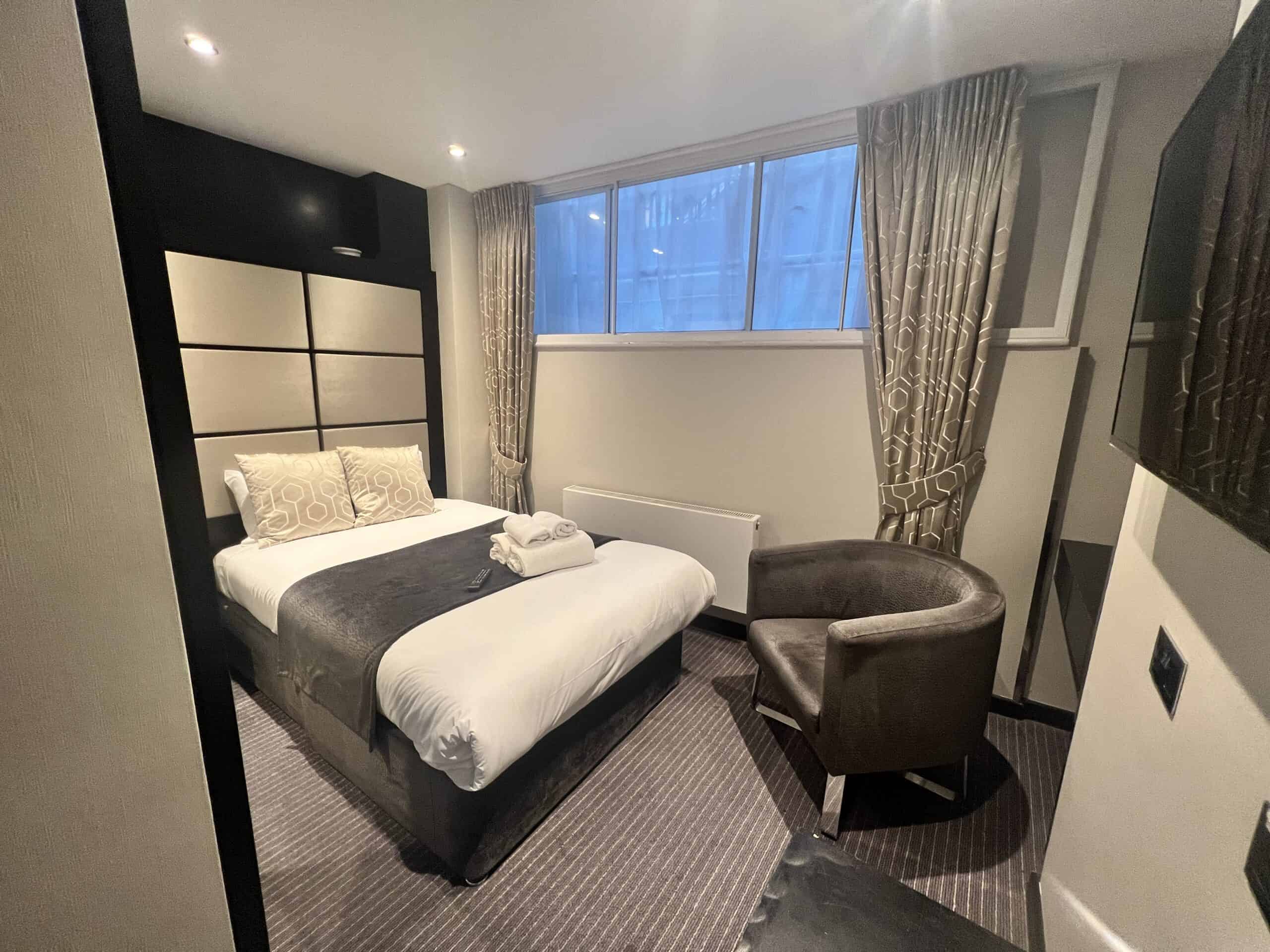 A cozy hotel room with a double bed featuring white and gray bedding. The room includes an armchair, folded towels on the bed, beige curtains, a wall-mounted flatscreen TV, and a window with frosted glass panels. The decor is modern with neutral tones.