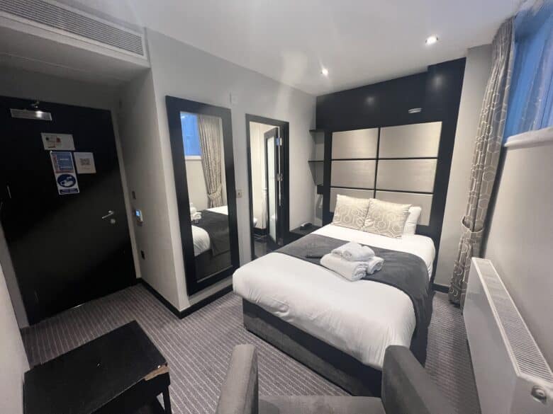 A modern hotel room features a neatly made double bed with white and gray bedding and accent pillows. A large mirror and a smaller mirror are on the wall beside the bed. Towels are folded on the bed, and a window with curtains allows natural light into the room.