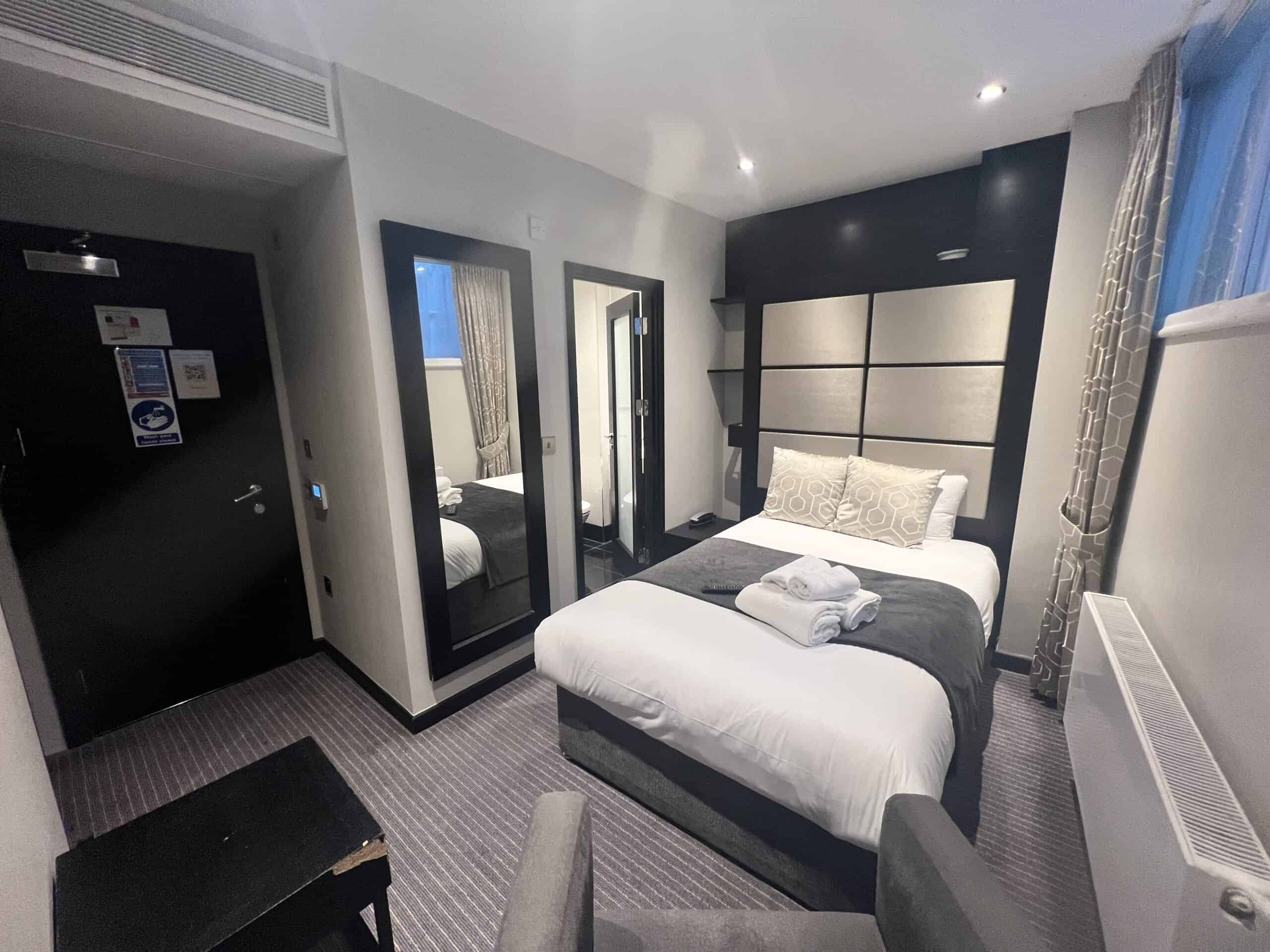 A modern hotel room features a neatly made double bed with white and gray bedding and accent pillows. A large mirror and a smaller mirror are on the wall beside the bed. Towels are folded on the bed, and a window with curtains allows natural light into the room.