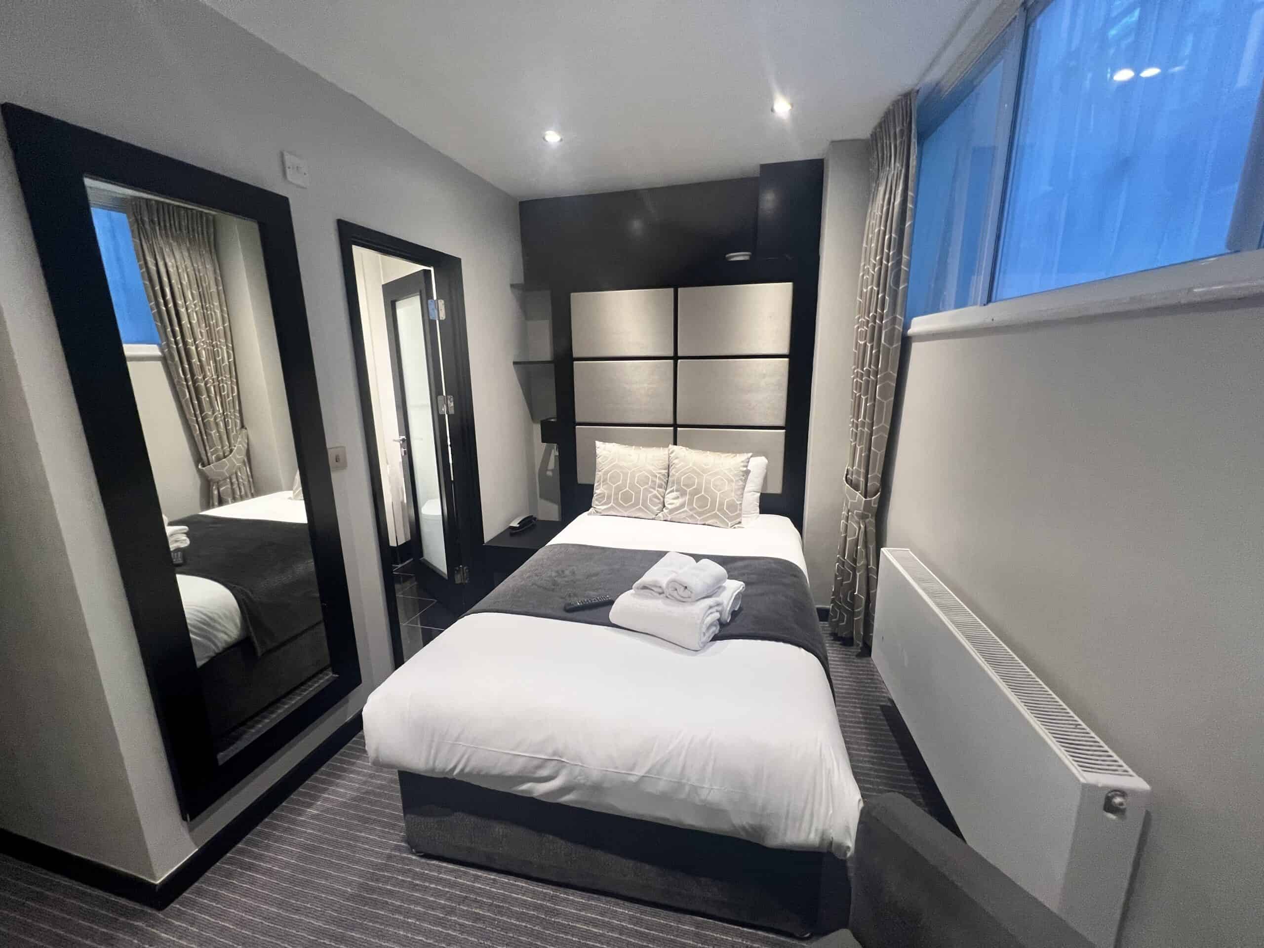 A small, modern bedroom with a single bed dressed in white and gray linens. Towels are neatly folded on the bed. The room features a large floor mirror, a radiator, and a window with gray curtains. The walls are adorned with a mix of light and dark panels.