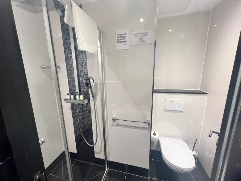 A modern bathroom with a walk-in shower enclosed by glass panels on the left, featuring a towel rack and toiletries. To the right is a toilet with a wall-mounted flush. The walls are tiled in white, and the floor has black tiles.