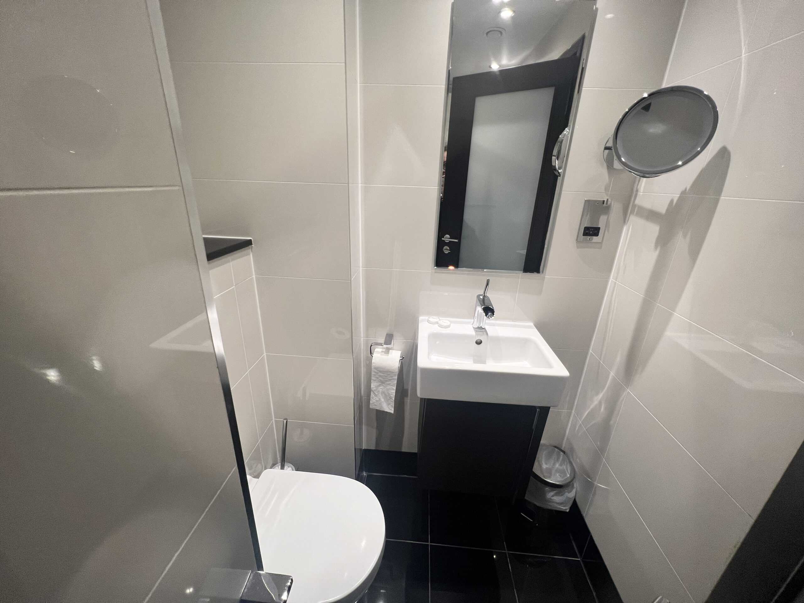 A modern bathroom with white tiled walls, a toilet, a small white sink with a mirror above it, a wall-mounted magnifying mirror, and a chrome toilet paper holder. The floor is covered with black tiles, and there is a small waste bin beside the sink.