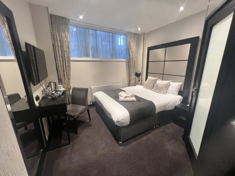 A modern hotel room with a large bed covered in white and gray linens, and two folded towels on the bed. There is a desk with a chair, a flat-screen TV mounted on the wall, and a large window with closed curtains. The room features minimalist decor.