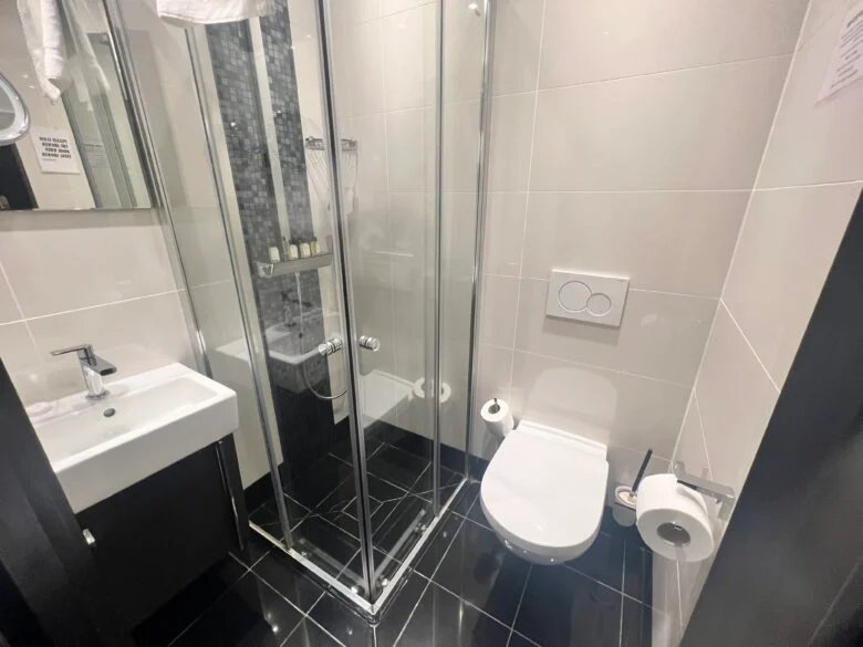 A compact, modern bathroom with white tiled walls and black tiled floors. It includes a glass-enclosed shower, a white sink with a mirror above it, and a wall-mounted toilet with toilet paper on a holder and a brush beside it. Various toiletries are visible in the shower.