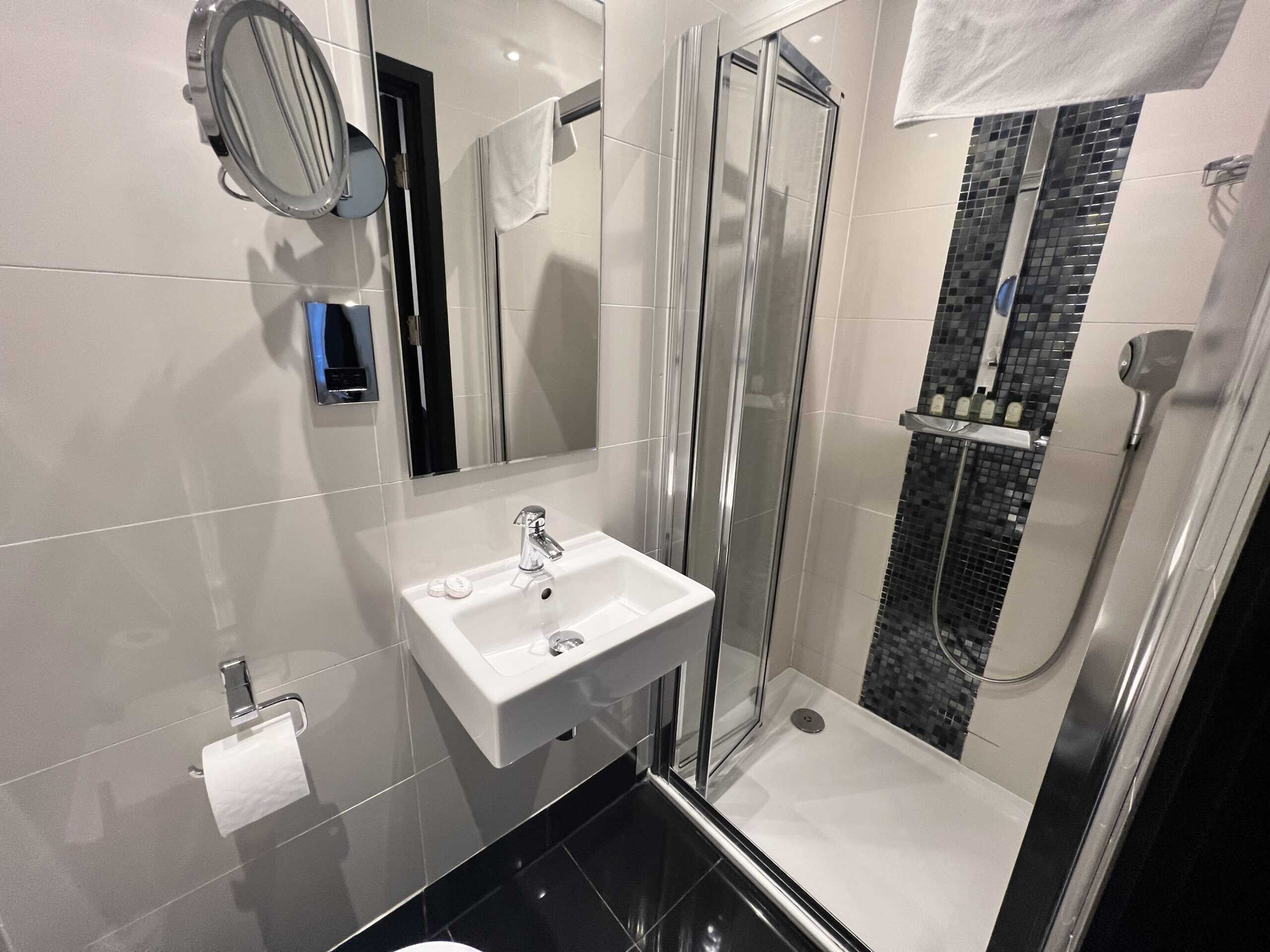 A modern bathroom with a white square sink and chrome fixtures is shown. A glass-enclosed shower with a black and white mosaic tile stripe is to the right, and a wall-mounted magnifying mirror and towel holder with a white towel are visible on the left.