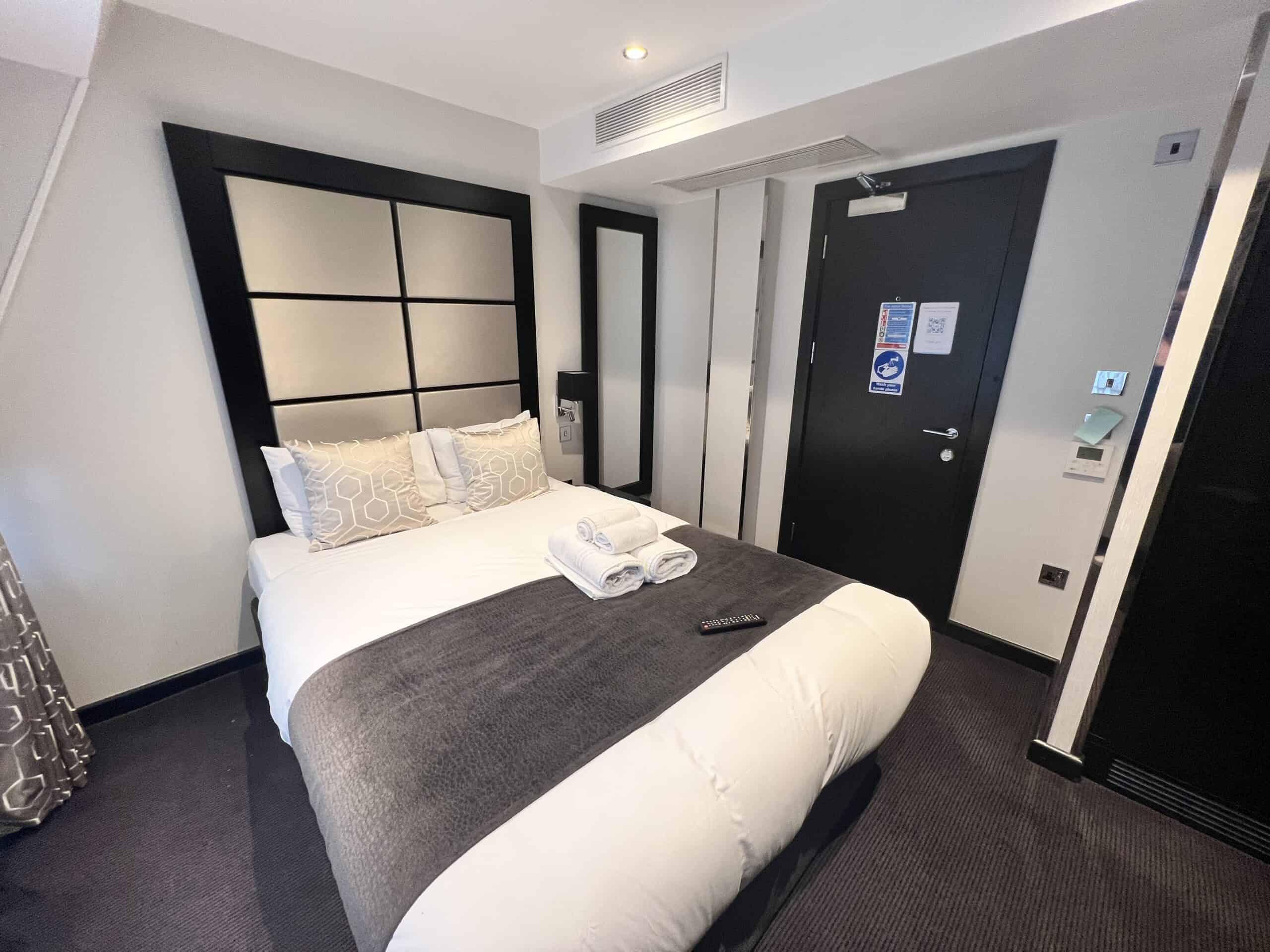 A modern hotel room featuring a neatly made double bed with white and gray bedding, and two folded towels on top. The room has a dark door with a small sign, a black headboard, gray carpet, and patterned curtains. A TV remote lies on the bed.