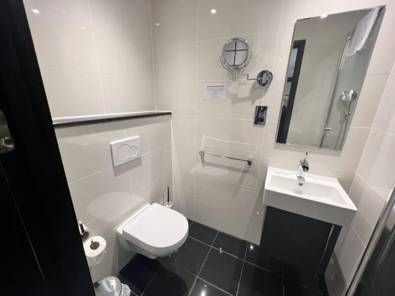 A compact bathroom with black-tiled flooring and white-tiled walls. It features a wall-mounted toilet, a small white rectangular sink with a mirror above it, a magnifying mirror on an extendable arm, and a stainless steel grab bar next to the toilet.