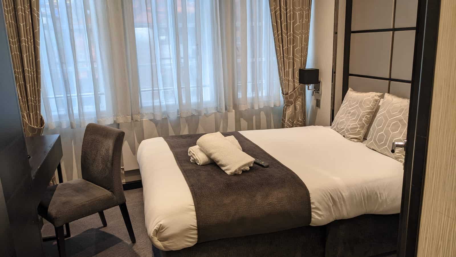 A cozy hotel room with a neatly made double bed, featuring brown and white bedding. There are rolled towels placed on the bed. A cushioned chair is positioned by a wooden desk against the left wall. The room is softly lit by natural light through sheer curtains.