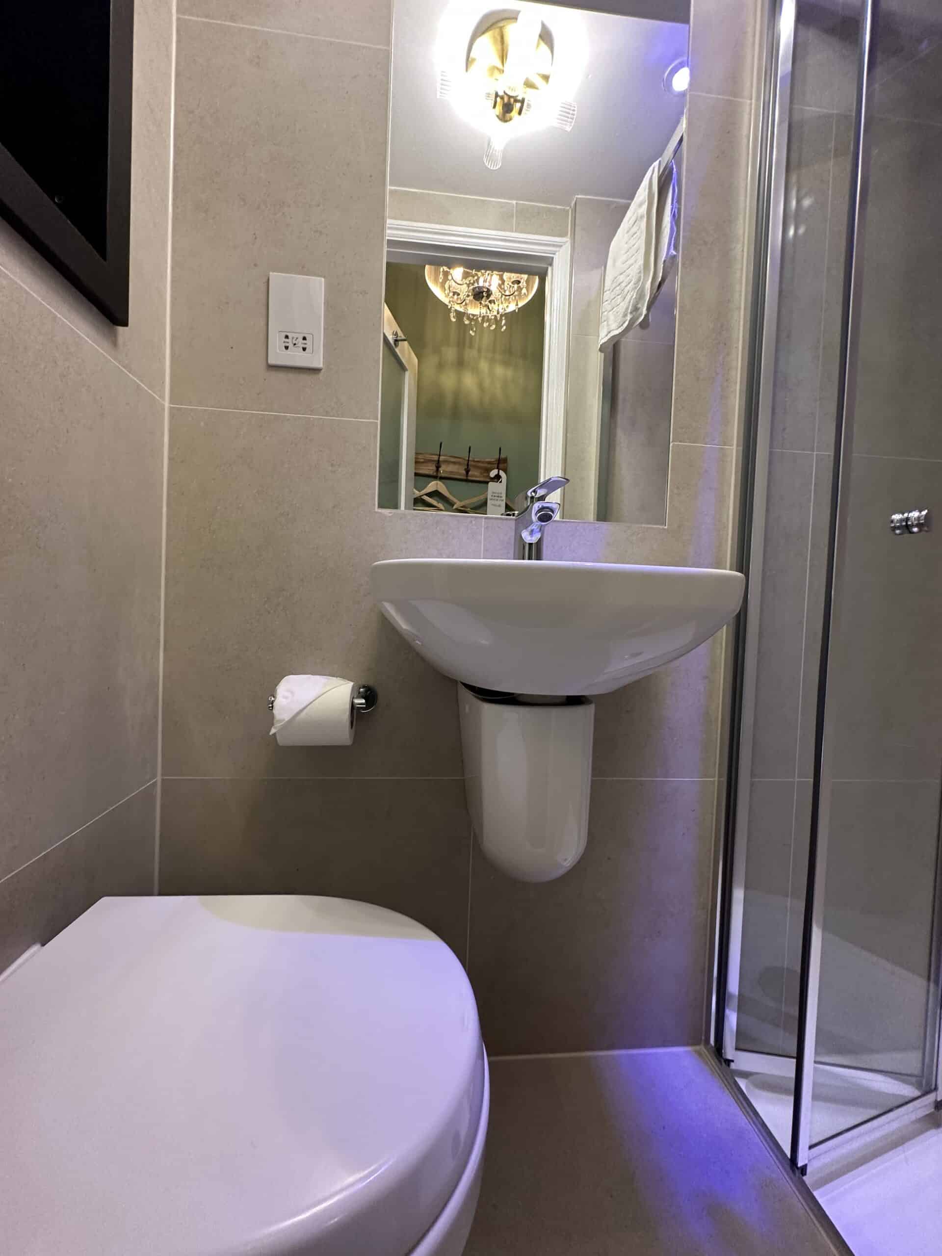 A compact bathroom with beige tiled walls features a white toilet, a wall-mounted sink with a faucet, a circular mirror, and a glass shower enclosure. A chandelier-style light fixture hangs from the ceiling, and a towel is draped over a rail.