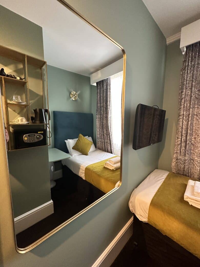 A small hotel room with a single bed adorned with green and white linens, reflected in a large wall mirror. There's a wall-mounted TV, a window with drapes, and a shelf containing a safe, tea kettle, mugs, and complimentary tea and coffee items.