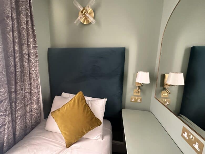 A small, neatly made bed with a white sheet and mustard yellow pillow leans against a padded, teal headboard. Above the headboard is a modern, brass wall sconce. A curved, wall-mounted mirror is on the right, beside matching sconces and power outlets.