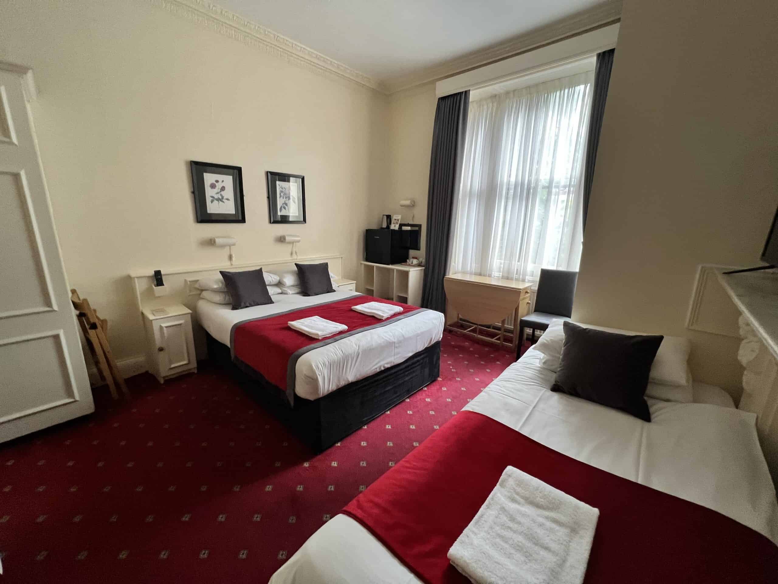 A cozy hotel room with two neatly made beds with red bed runners and white sheets. The room features a red carpet, a large window with curtains, a desk with a chair by the window, framed artwork on the walls, and a small nightstand with a lamp.