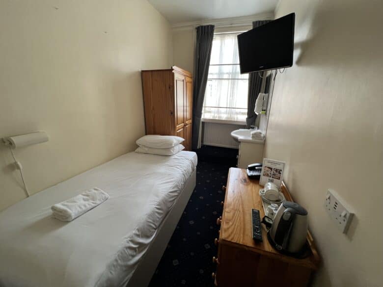 Compact hotel room with a single bed, wooden wardrobe, and a small desk holding a tea kettle, cups, and a TV remote. A wall-mounted flat-screen TV is angled towards the bed. A window with curtains lets in natural light, and a towel is placed on the bed.