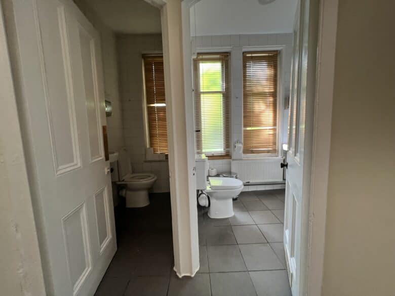 A view through an open door into a bathroom with large windows covered by wooden blinds. The room features a white toilet, a sink, and white tiled walls. The floor is covered with grey tiles.