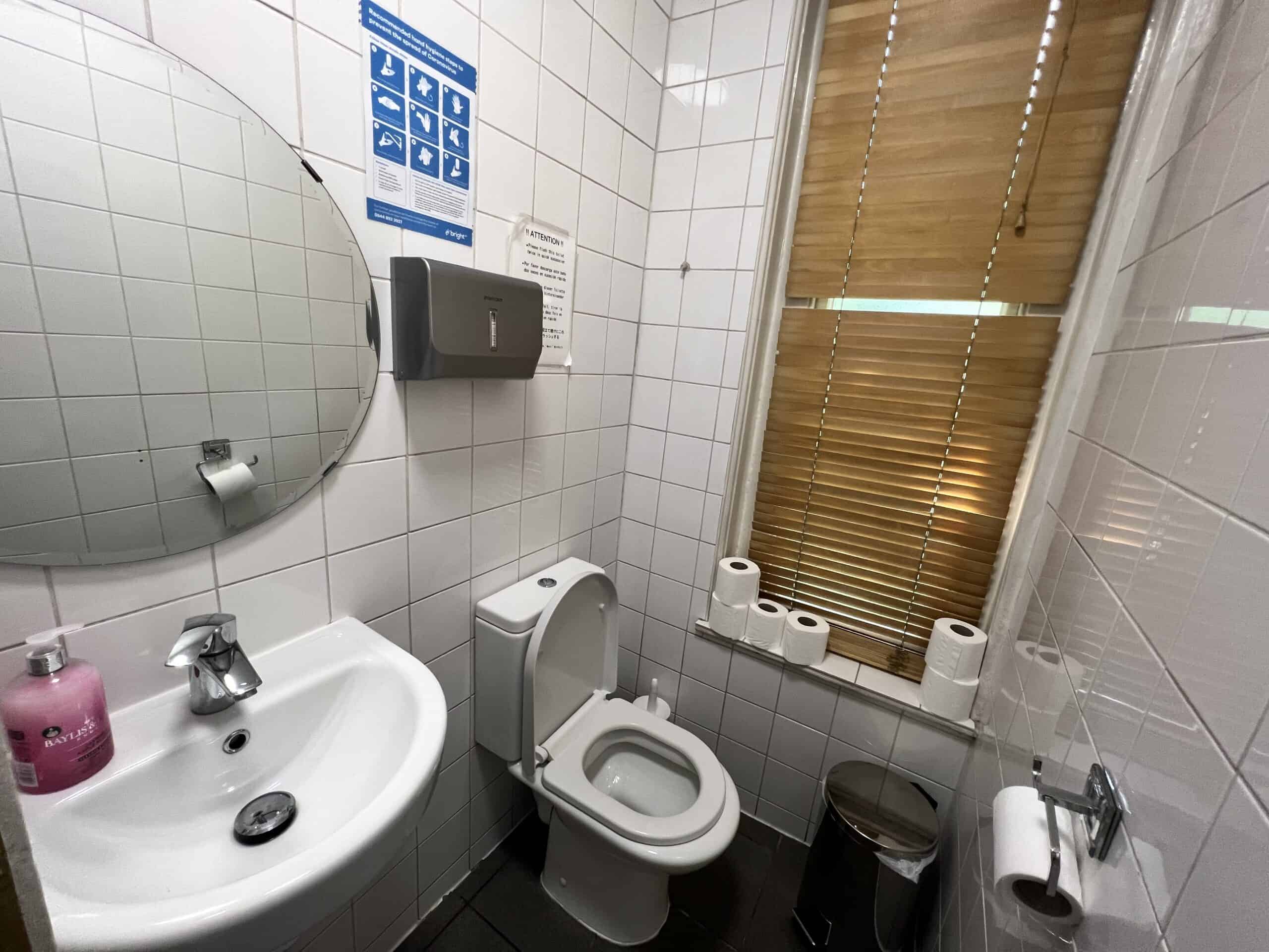 A small bathroom with white tiled walls featuring a round mirror above a white sink with a pink soap dispenser, a wall-mounted paper towel dispenser, a toilet, toilet paper rolls on a windowsill, and a window with closed blinds.