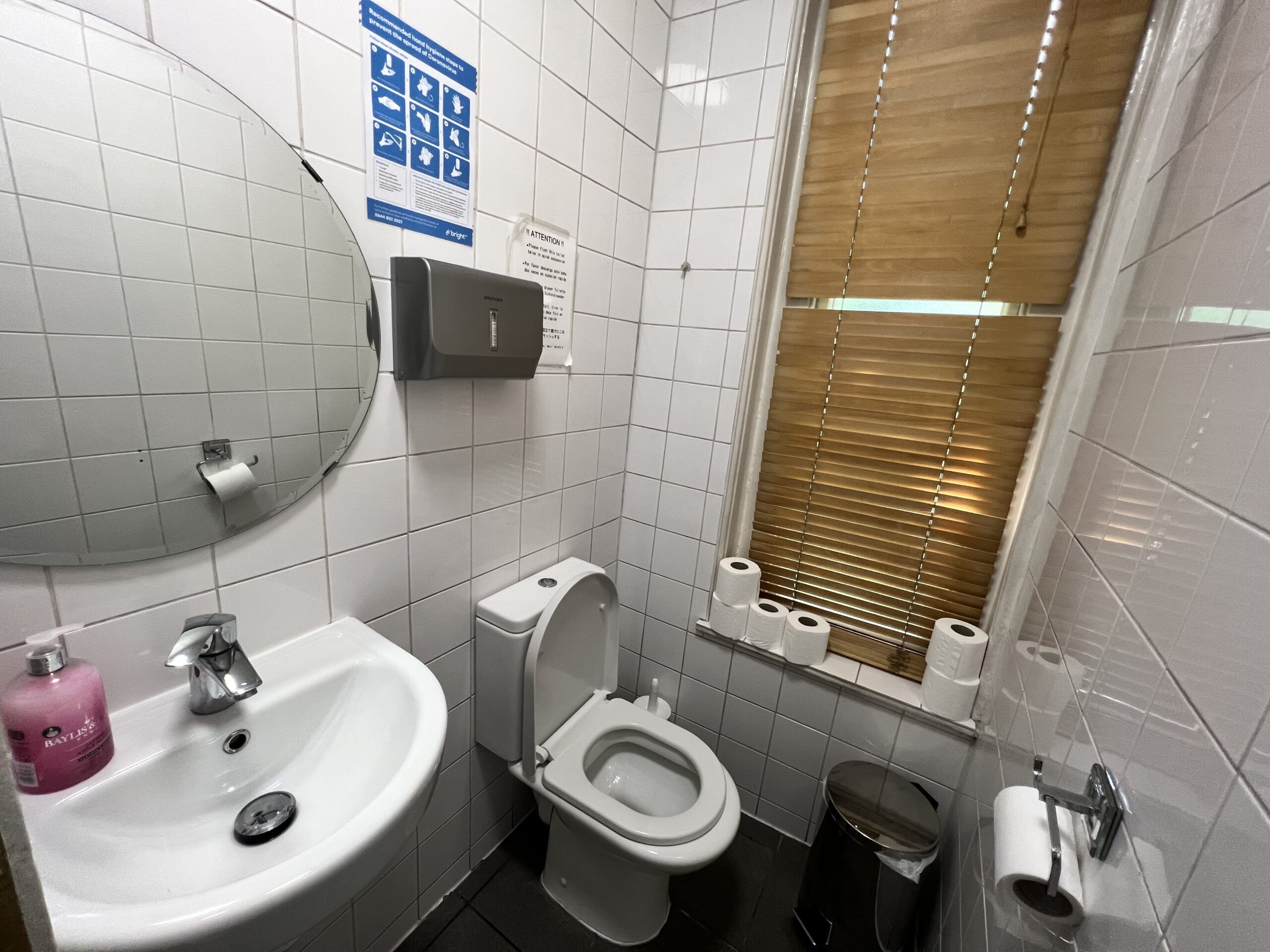 A small, clean bathroom with white-tiled walls featuring a round mirror above a white sink with a soap dispenser, a wall-mounted paper towel dispenser, a white toilet, several rolls of toilet paper on a windowsill, and a silver trash can.